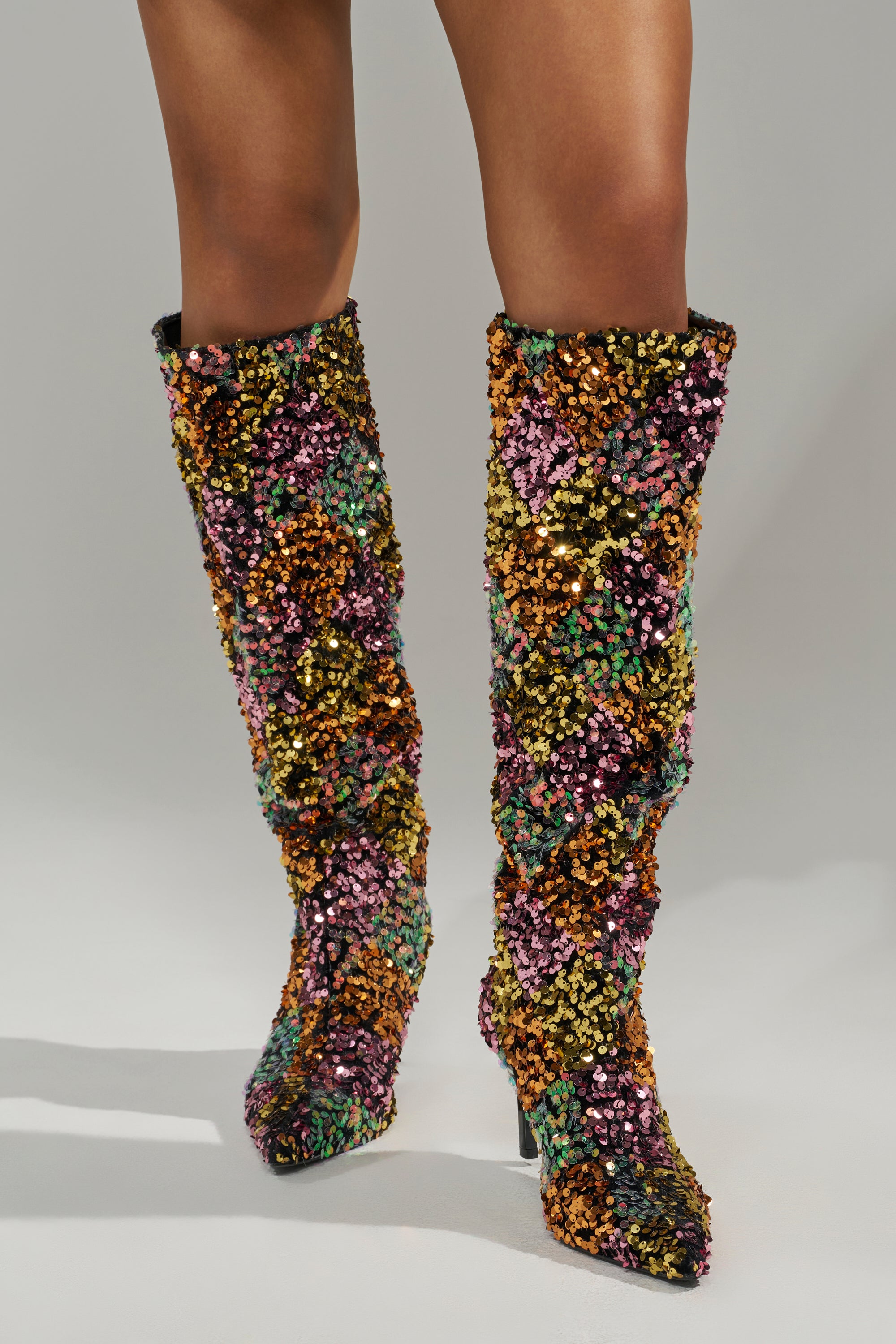 A person models only the lower legs and AZALEA WANG JIMENA MULTI GOLD SEQUIN KNEE HIGH STILETTO BOOTS, covered in gold, purple, green, and orange sequins, against a plain light background.