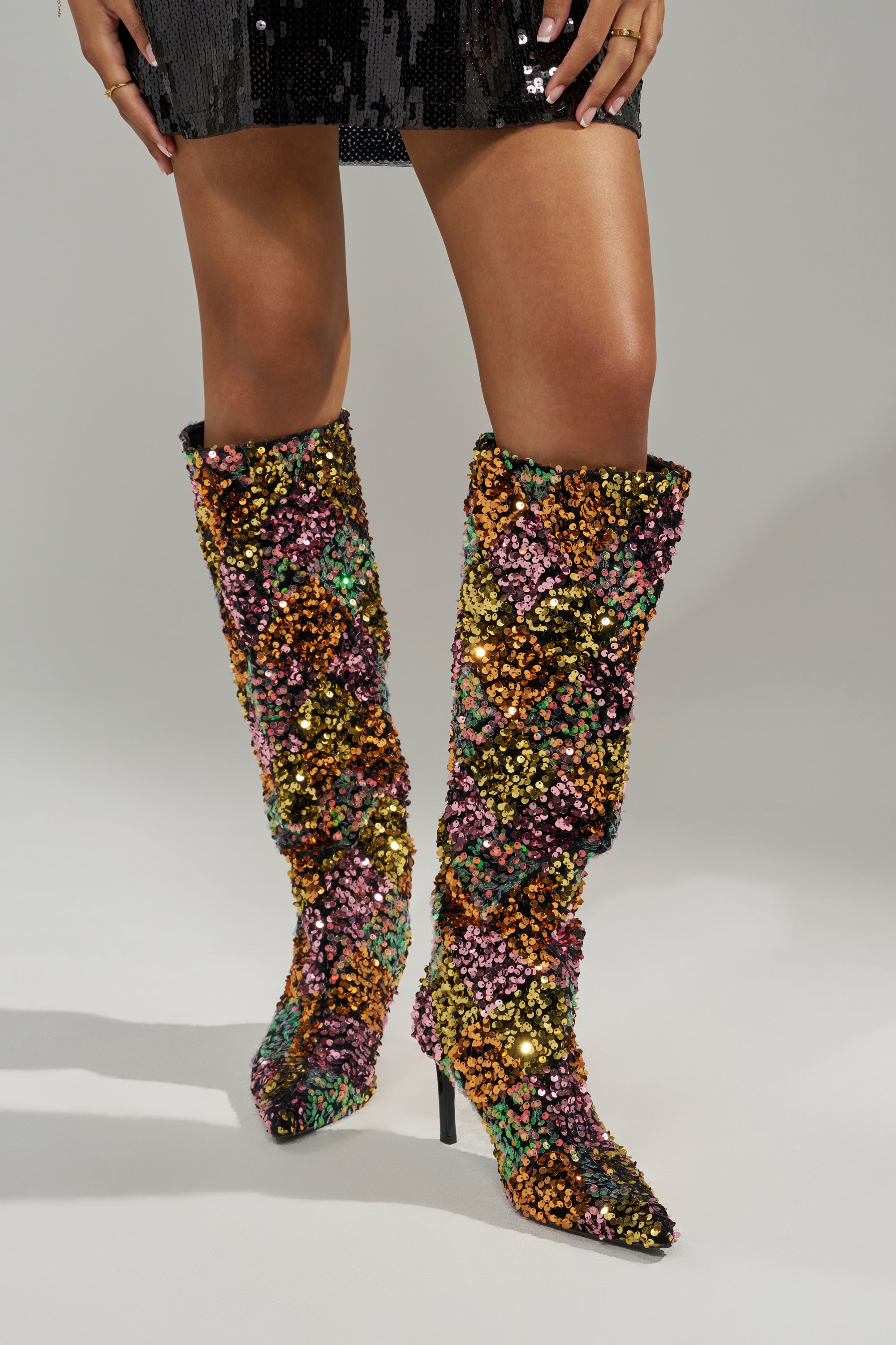 Someone stands against a plain background wearing a black sequin mini skirt and AZALEA WANG JIMENA MULTI GOLD SEQUIN KNEE HIGH STILETTO BOOTS in shimmering pink, green, and purple.