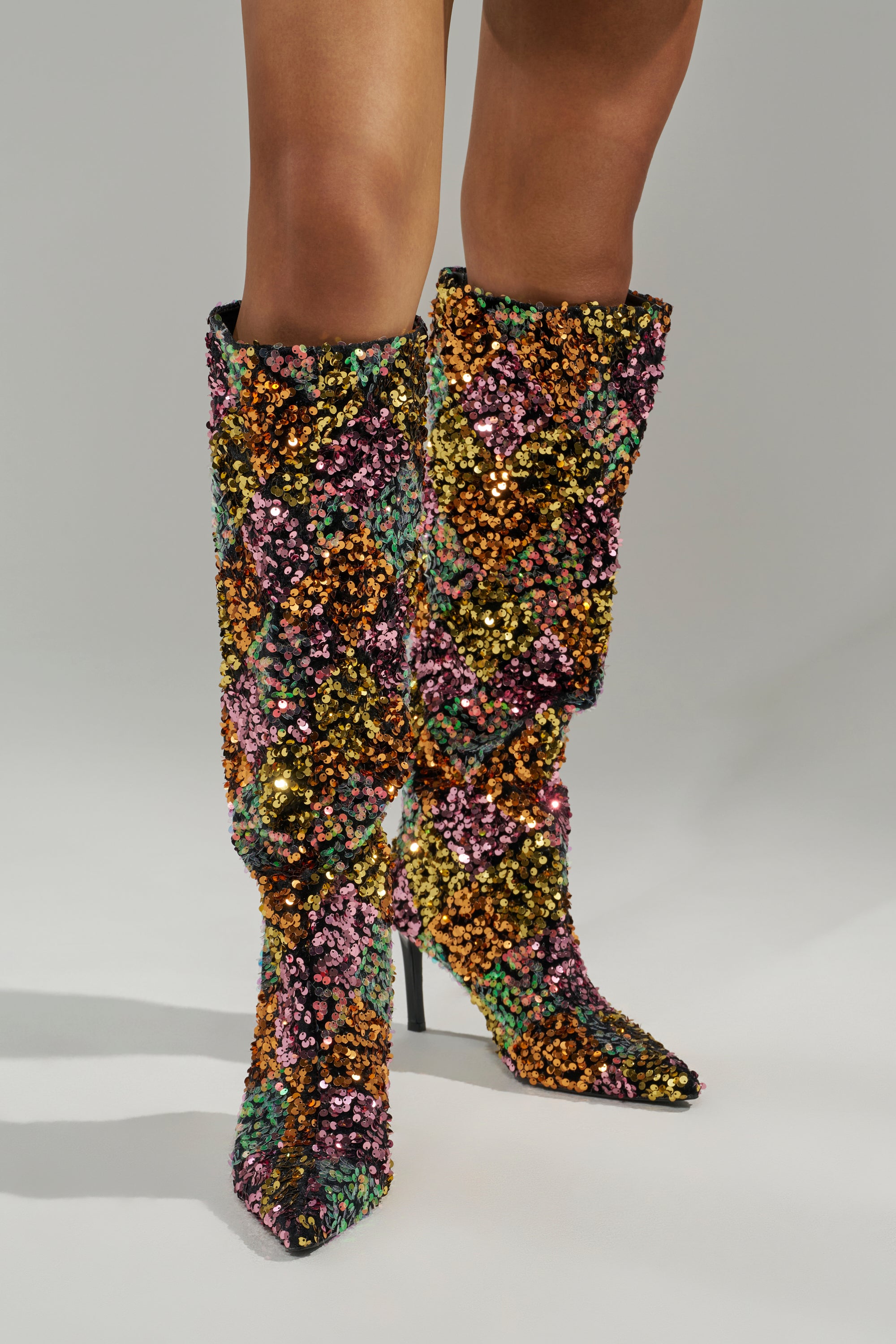 A person wears the AZALEA WANG JIMENA MULTI GOLD SEQUIN KNEE HIGH STILETTO BOOT, adorned with multicolored sequins, while standing against a plain gray background.