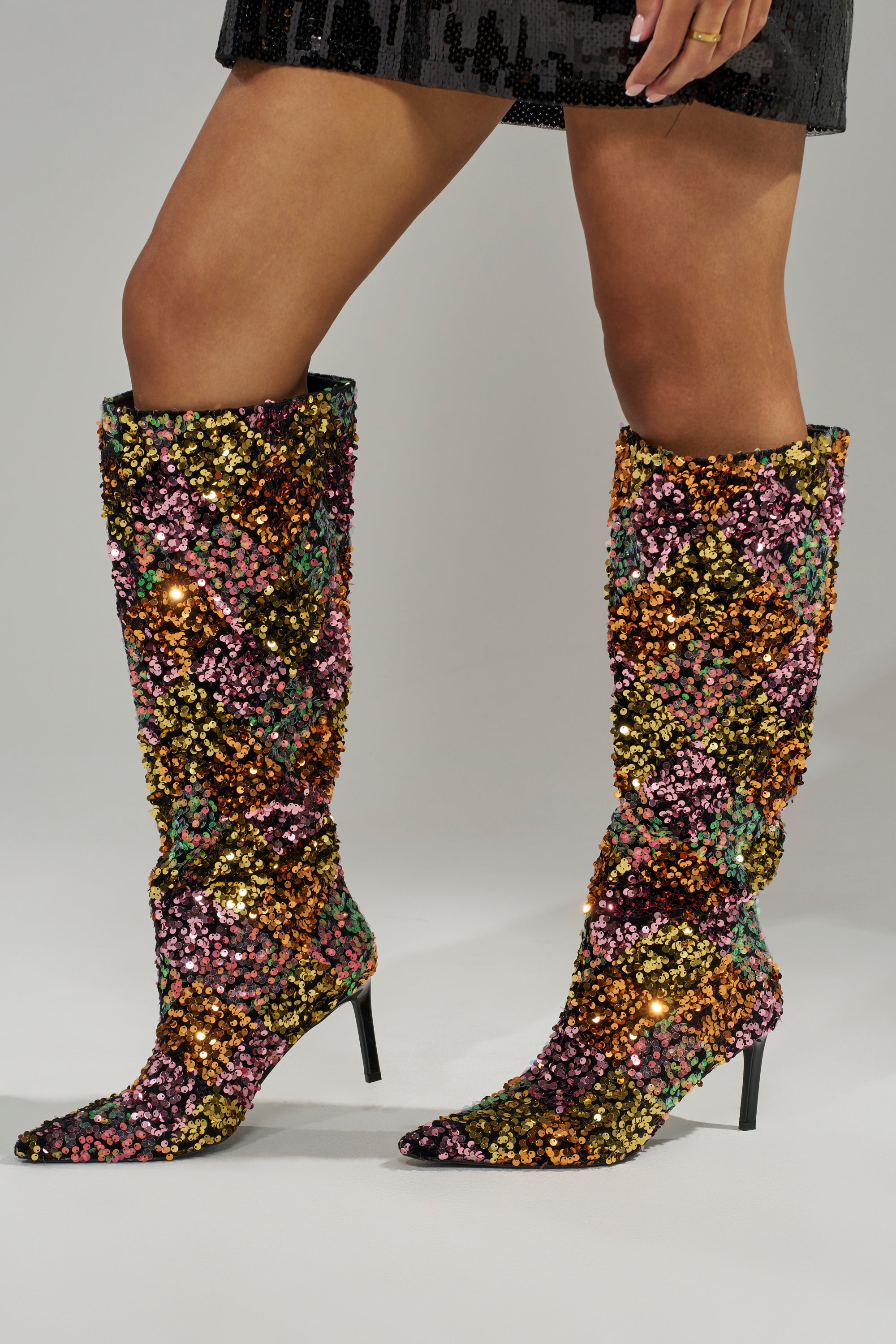 A person wears the AZALEA WANG JIMENA MULTI GOLD SEQUIN KNEE HIGH STILETTO BOOT, featuring pointed toes and sparkling multicolored sequins, paired with a black sequin skirt for a chic, vibrant look.