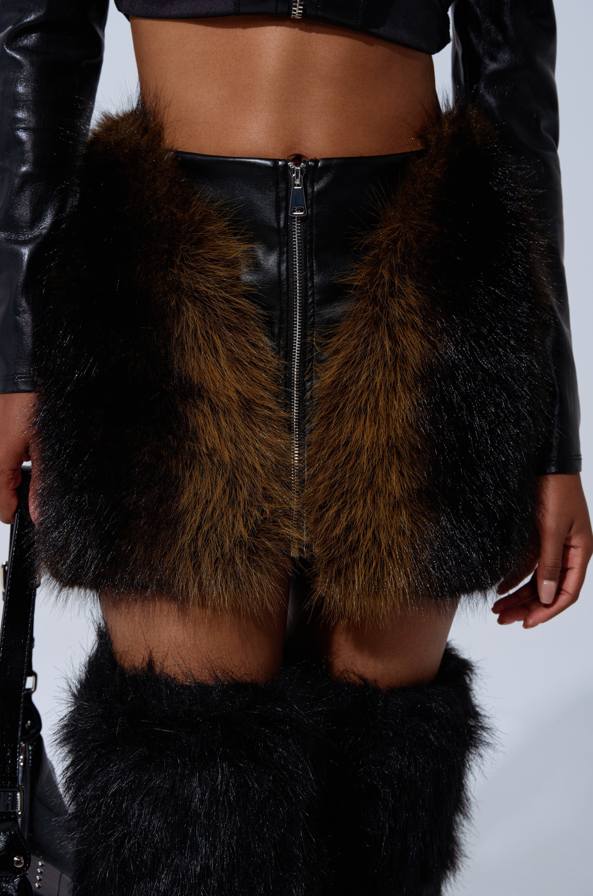 A person is wearing the TOUCH ME IN THE MORNING FUR SKIRT, a high-rise mini with brown and black faux fur panels, paired with a black leather crop top and matching black furry leg warmers. Only the torso and legs are visible. 1