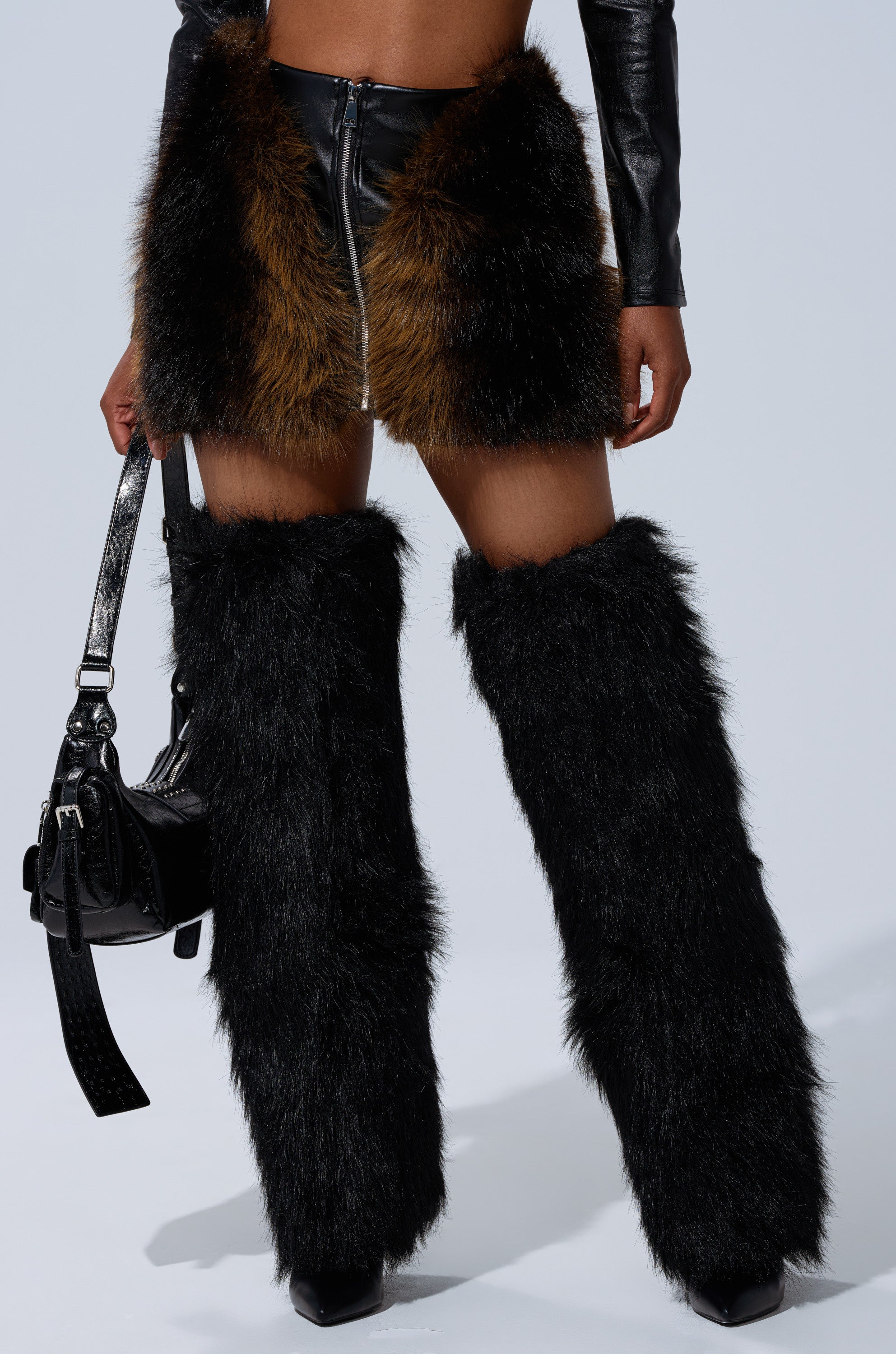 A person styles the TOUCH ME IN THE MORNING FUR SKIRT, pairing its black faux leather and brown fur panels with black furry leg warmers, high-heeled boots, and a black handbag for a bold, textured, modern look. 2