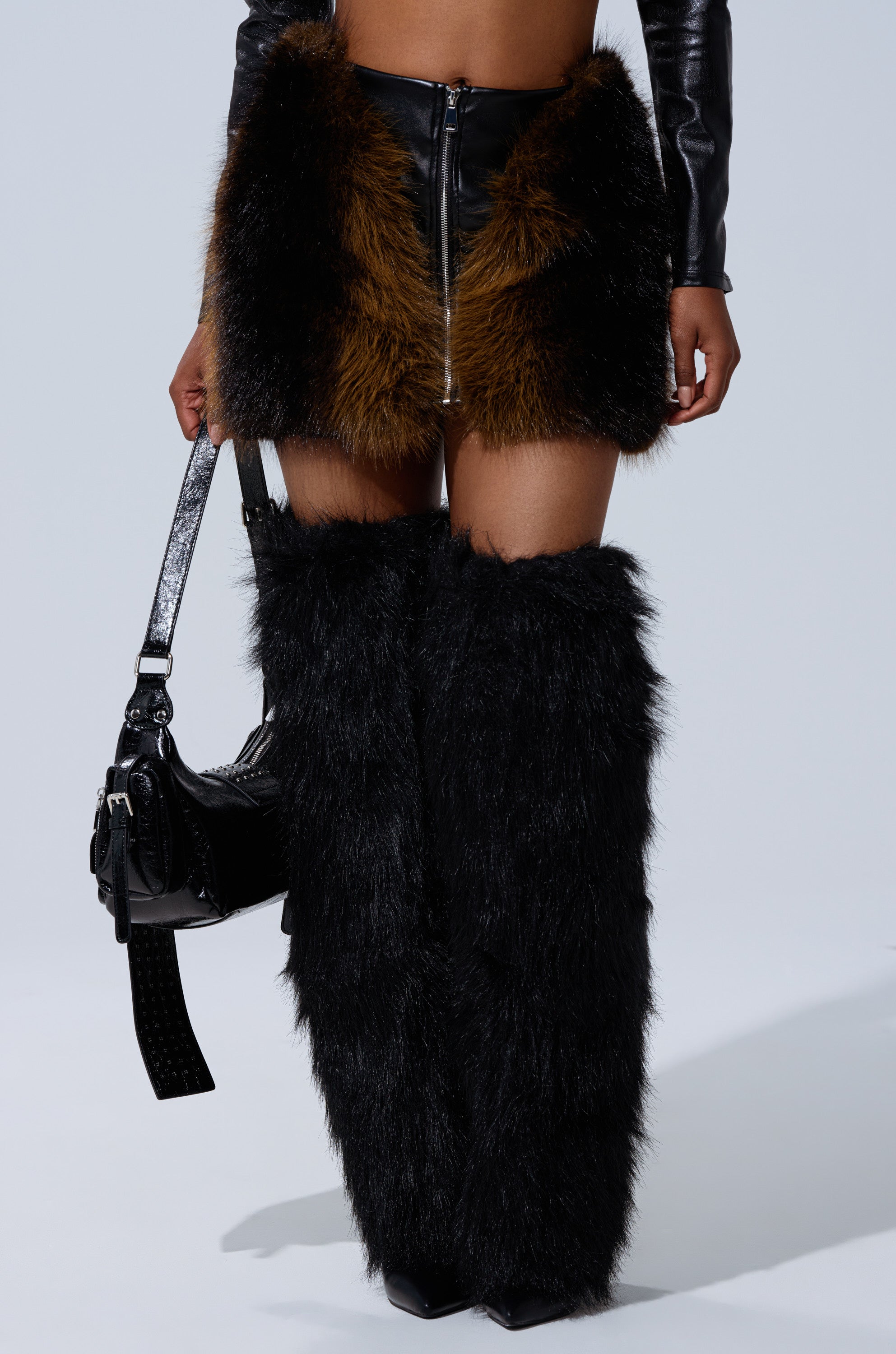 A person stands against a light background wearing the TOUCH ME IN THE MORNING FUR SKIRT, long black furry boots, a black leather purse, and a black long-sleeve top. 3