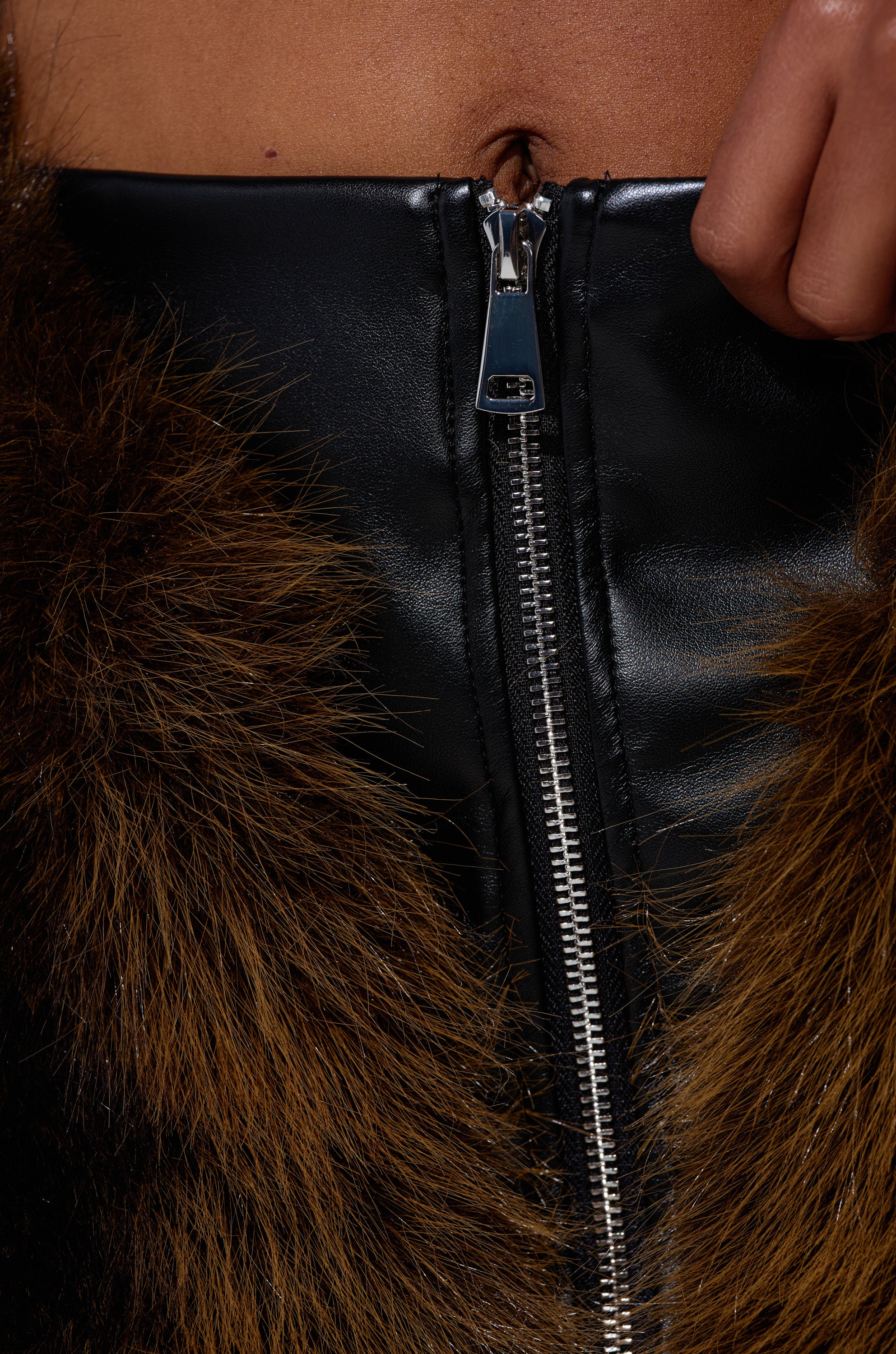 Close-up of a person's torso wearing the TOUCH ME IN THE MORNING FUR SKIRT, a black faux leather skirt with zipper and brown faux fur trim. The person holds the skirt by the zipper, revealing their belly button. 7