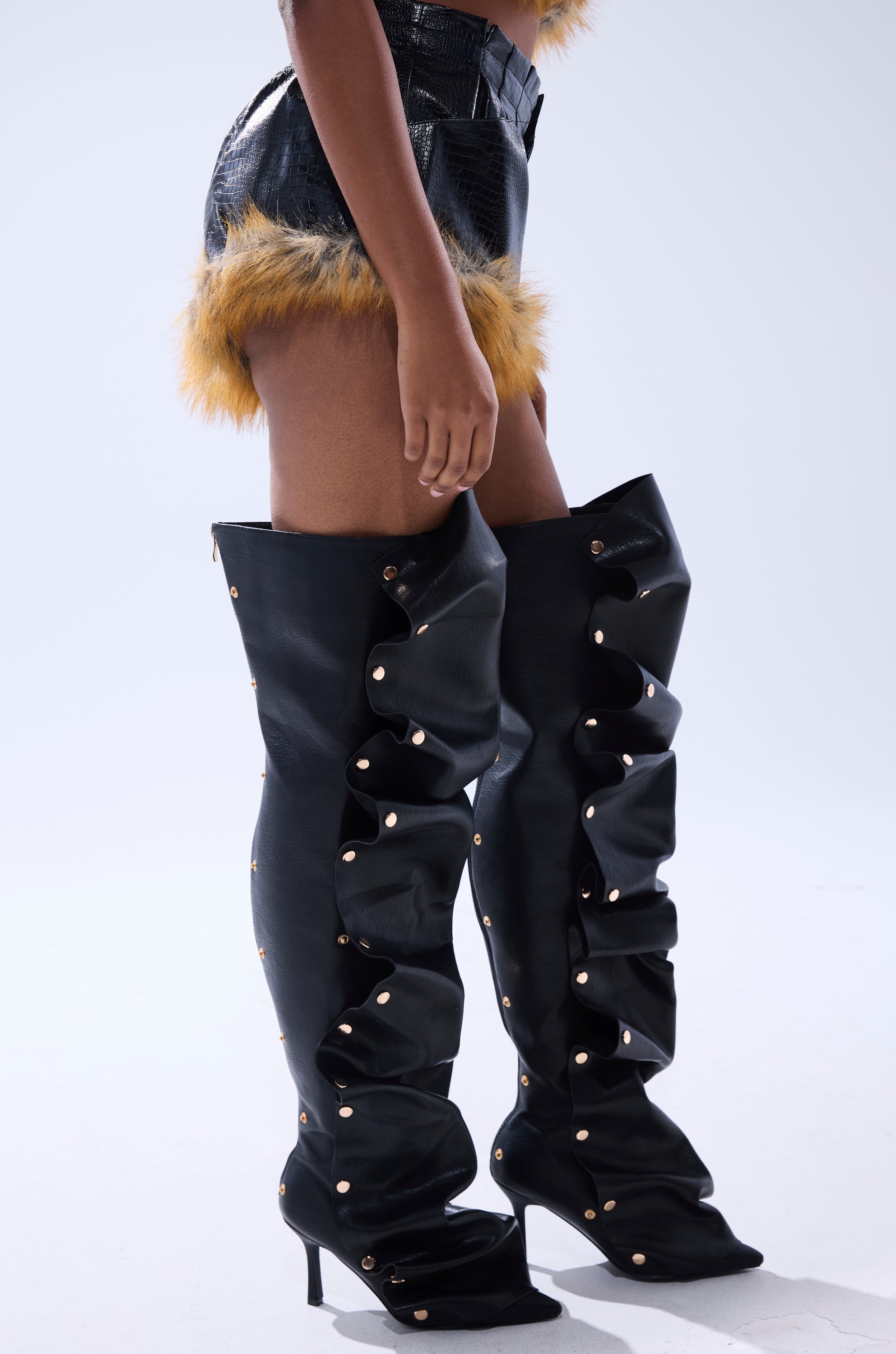 Wearing the DONT TOUCH OR LOOK FAUX LEATHER SHORT WITH FUR TRIM, a person stands against a plain light background in black crocodile-embossed faux leather shorts with yellow fur trim and ruffled, studded thigh-high boots.