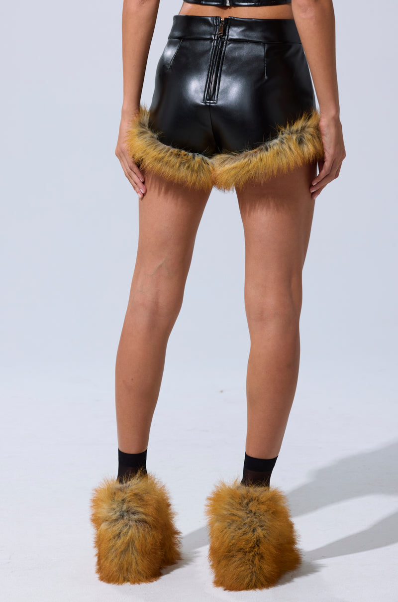 A person faces away wearing DONT TOUCH SHORT WITH FUR TRIM—shiny black high-rise faux leather shorts with tan faux fur trim—and matching tan faux fur leg warmers over black socks, against a plain light background. 5