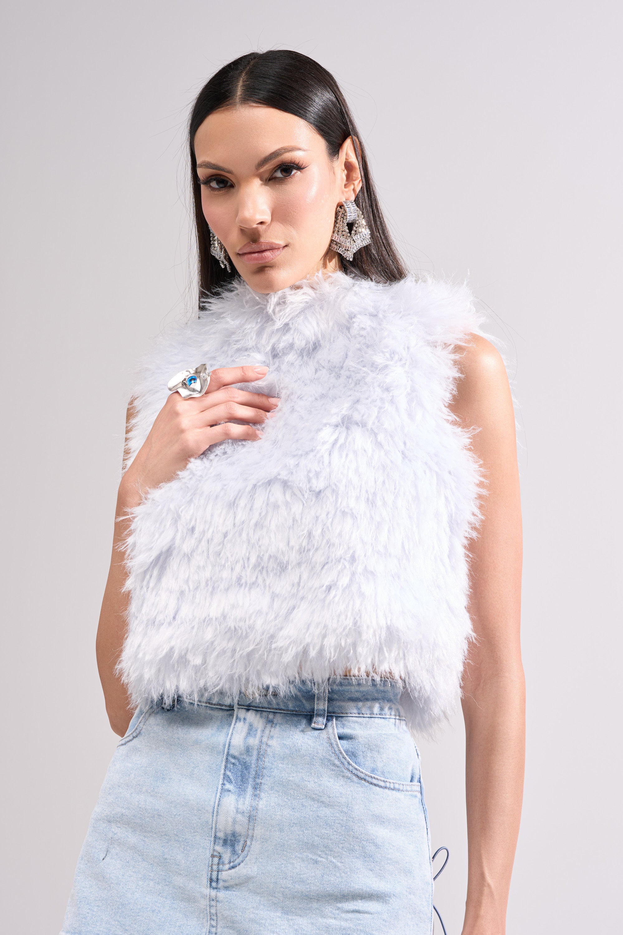A woman models the ICY OPEN BACK FUR TOP, a sleeveless cropped faux fur piece, paired with high-waisted light blue jeans. She has long straight dark hair and bold accessories, posing against a plain light background.