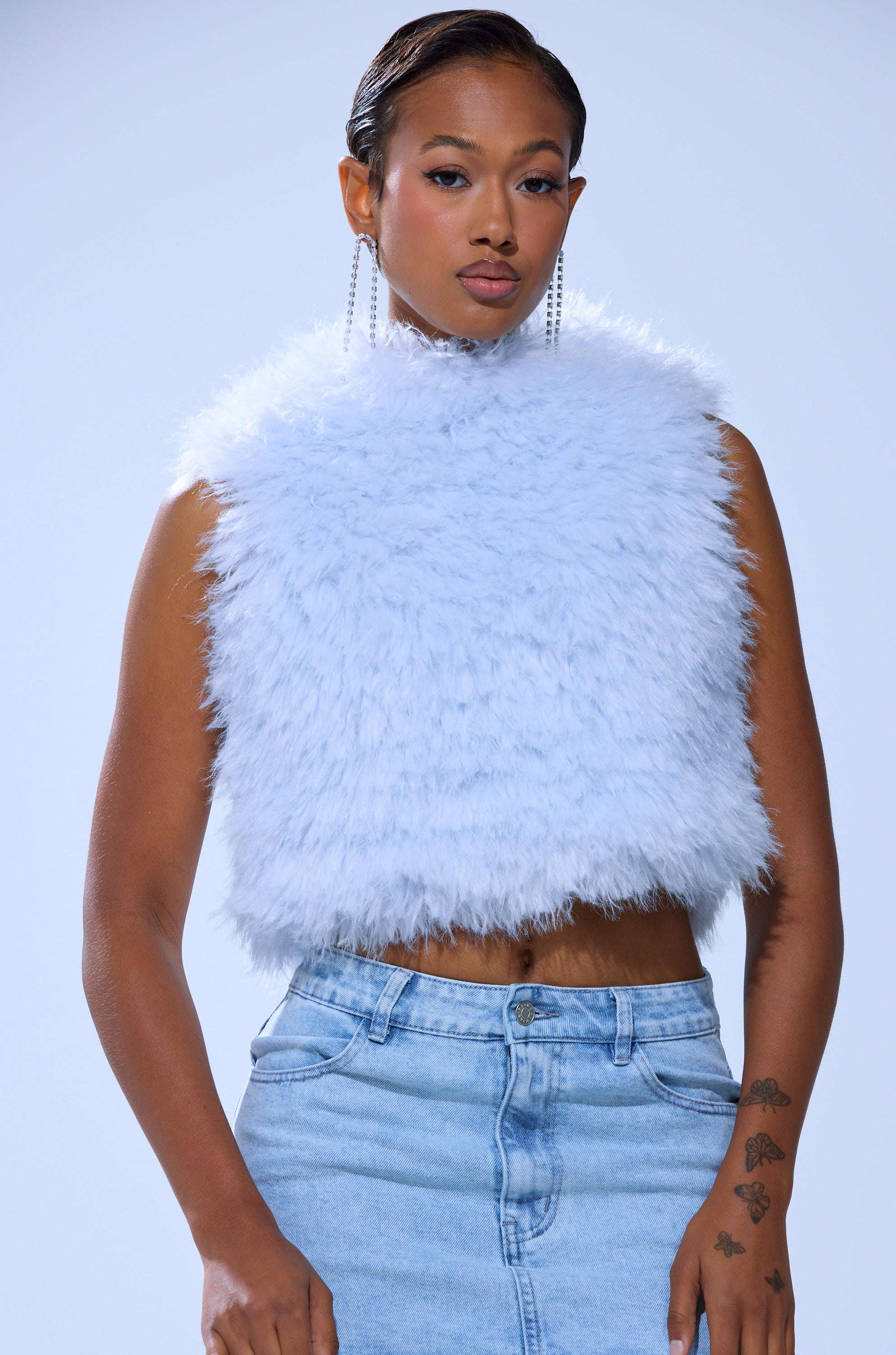 A woman poses against a plain background wearing the ICY OPEN BACK FUR TOP, a sleeveless, cropped light blue faux fur piece, paired with high-waisted jeans. She has slicked-back hair, long earrings, and hand tattoos.