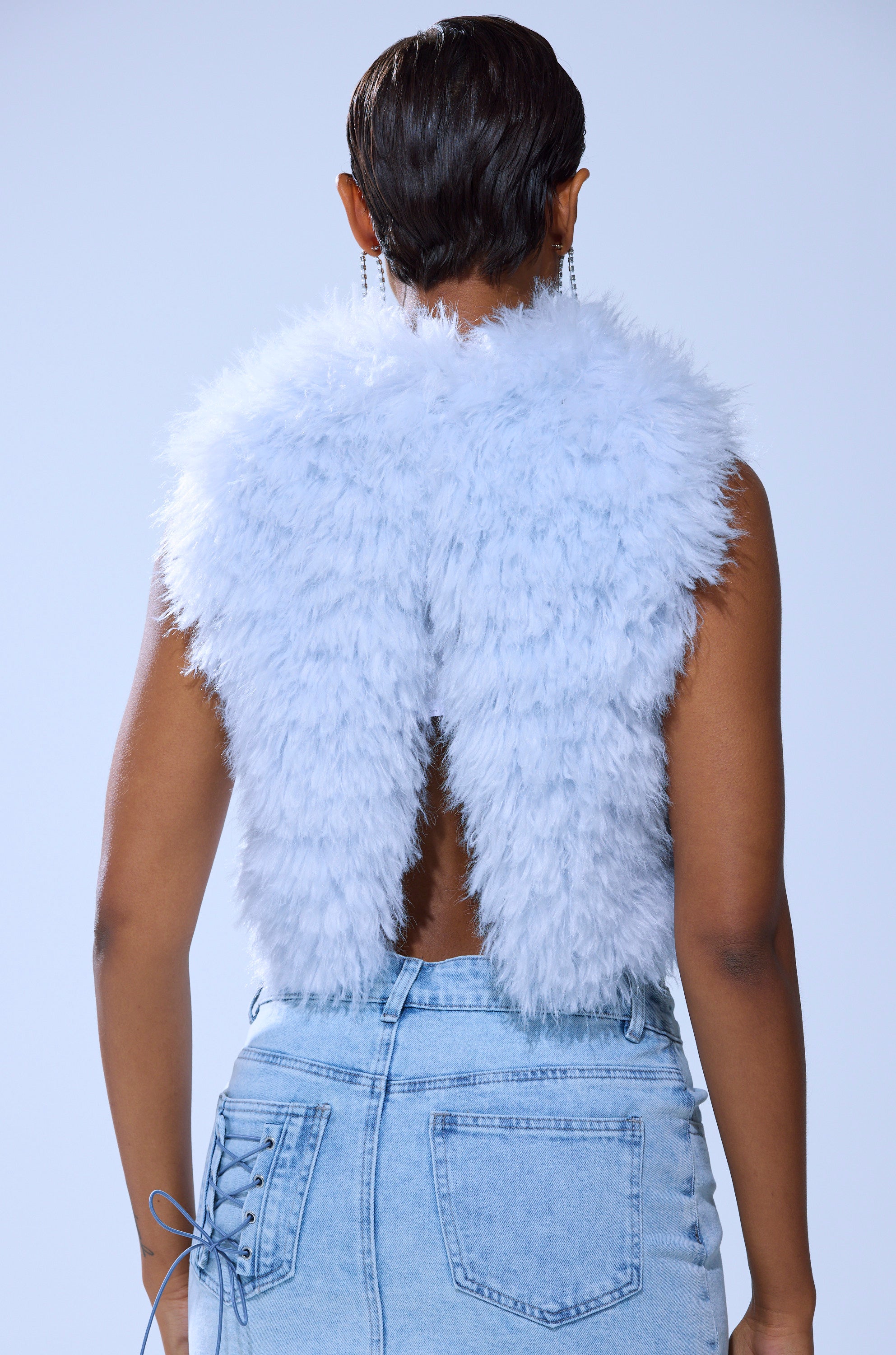 A person with short hair and dangling earrings wears the ICY OPEN BACK FUR TOP in light blue, styled with a light wash denim skirt featuring side lace-up details.