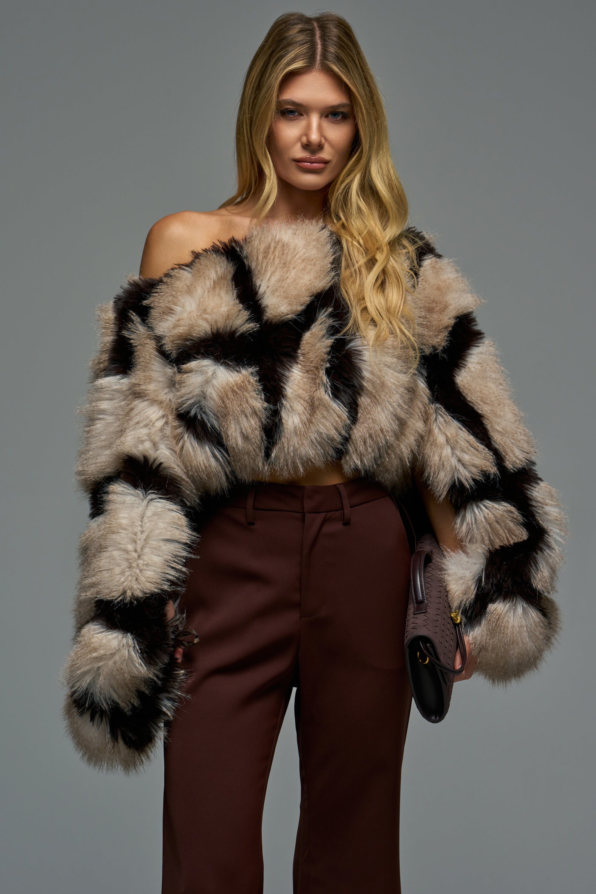 A woman wears the SNOW DAY FAUX FUR SWEATER IN BROWN MULTI, an off-the-shoulder faux fur sweater with a tan and black abstract checkerboard pattern, paired with dark brown trousers.