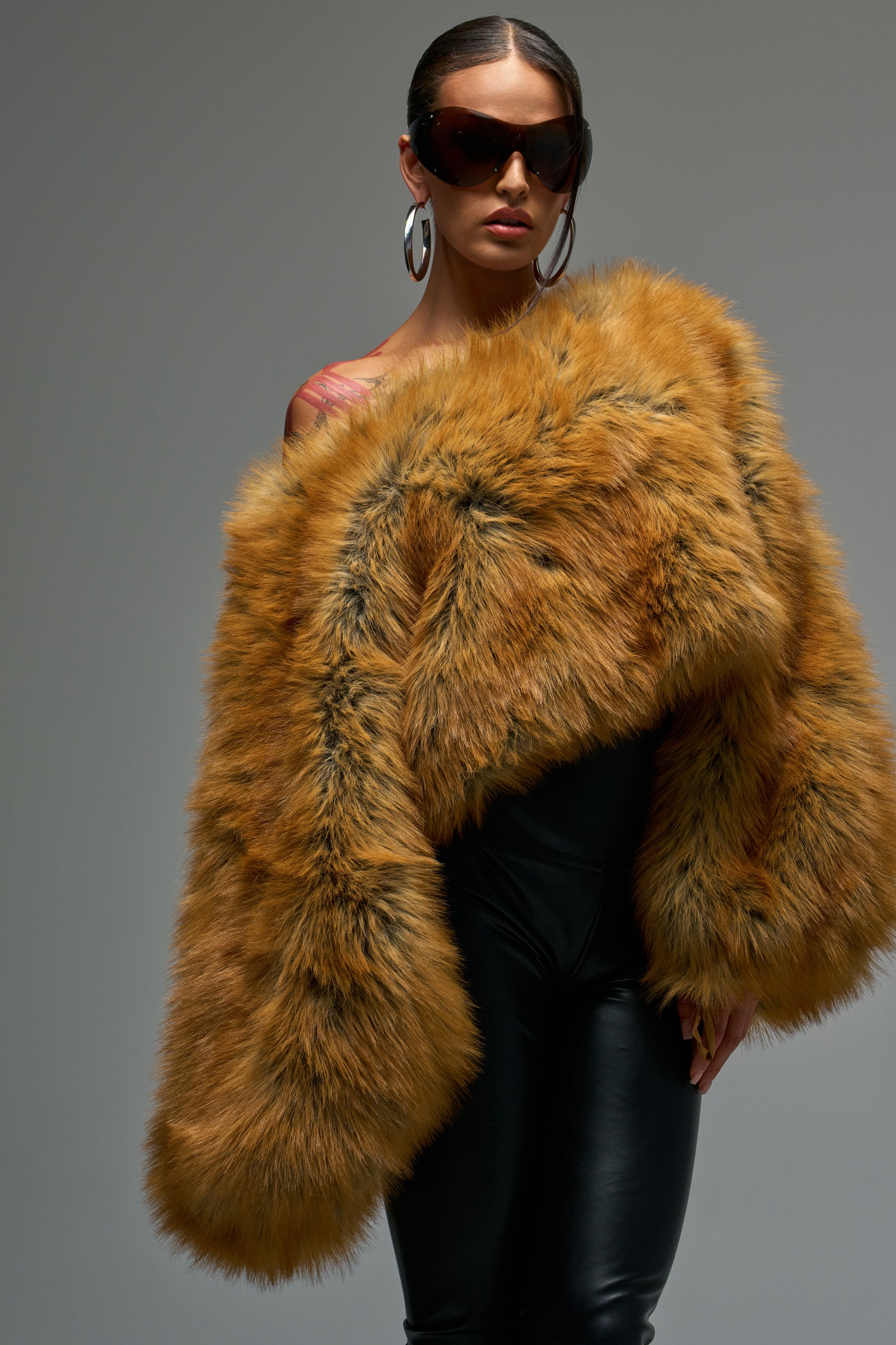 A woman poses confidently in après-chic cold weather style, wearing the SNOW DAY FAUX FUR SWEATER draped off one shoulder with black leather pants, large sunglasses, and silver hoop earrings against a neutral background.