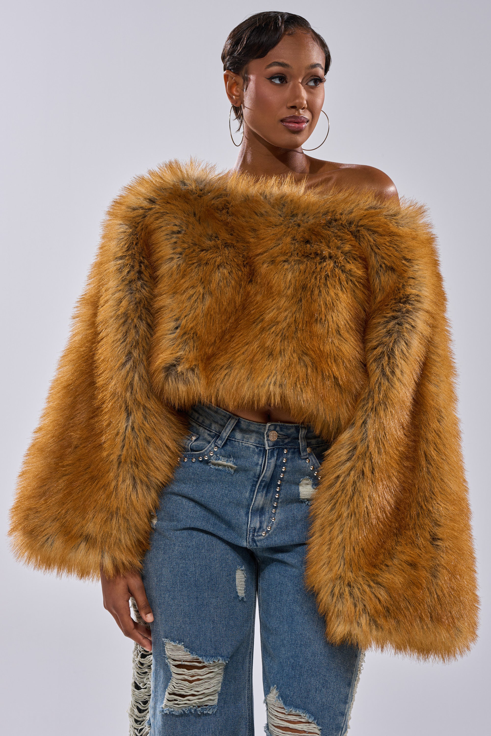 A woman wears the bold orange SNOW DAY FAUX FUR SWEATER off the shoulder, paired with high-waisted ripped jeans, standing against a plain background.