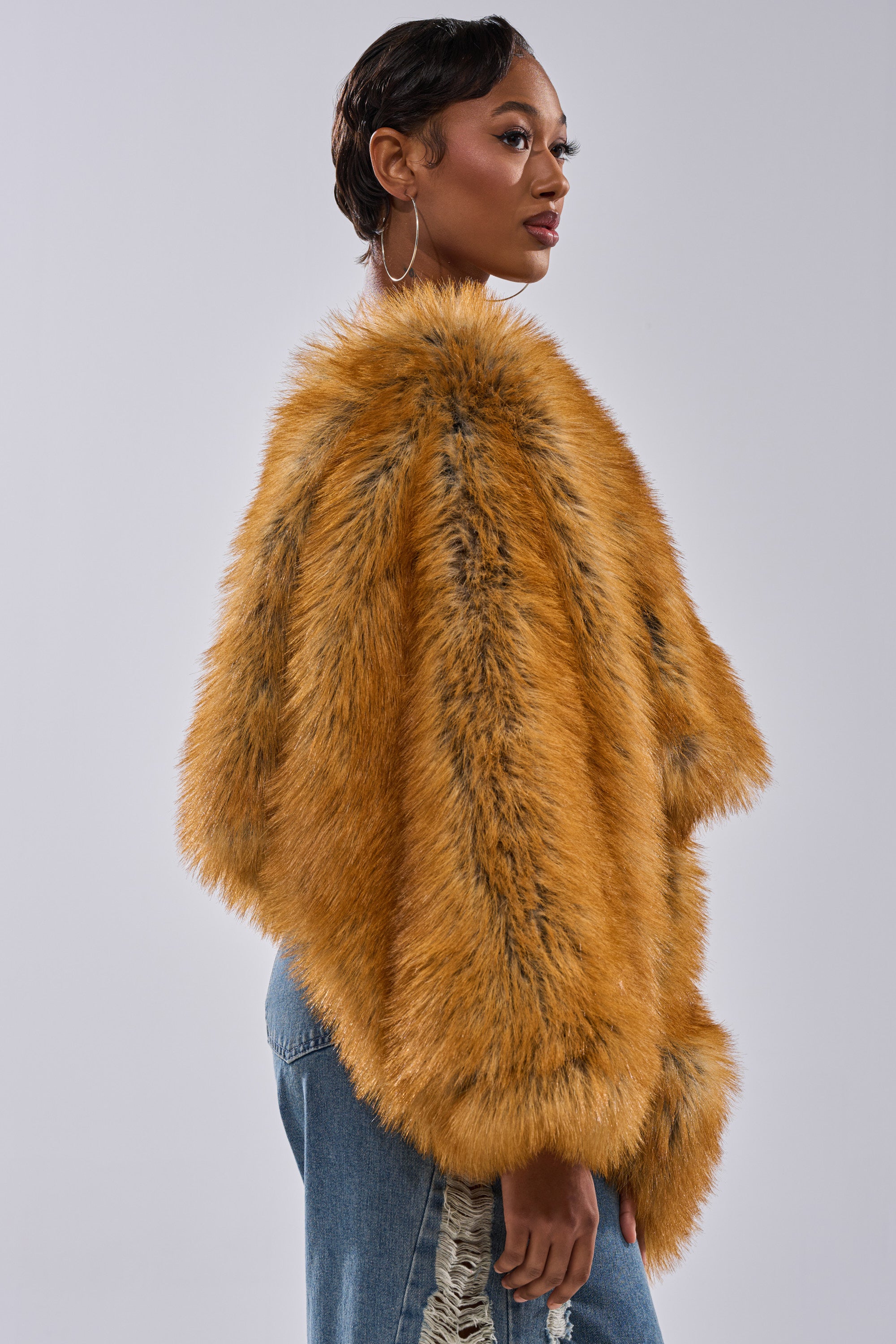 A woman stands sideways wearing the SNOW DAY FAUX FUR SWEATER, a golden brown faux fur coat with black streaks, paired with blue jeans. She has short, styled hair and large hoop earrings, set against a plain light background.
