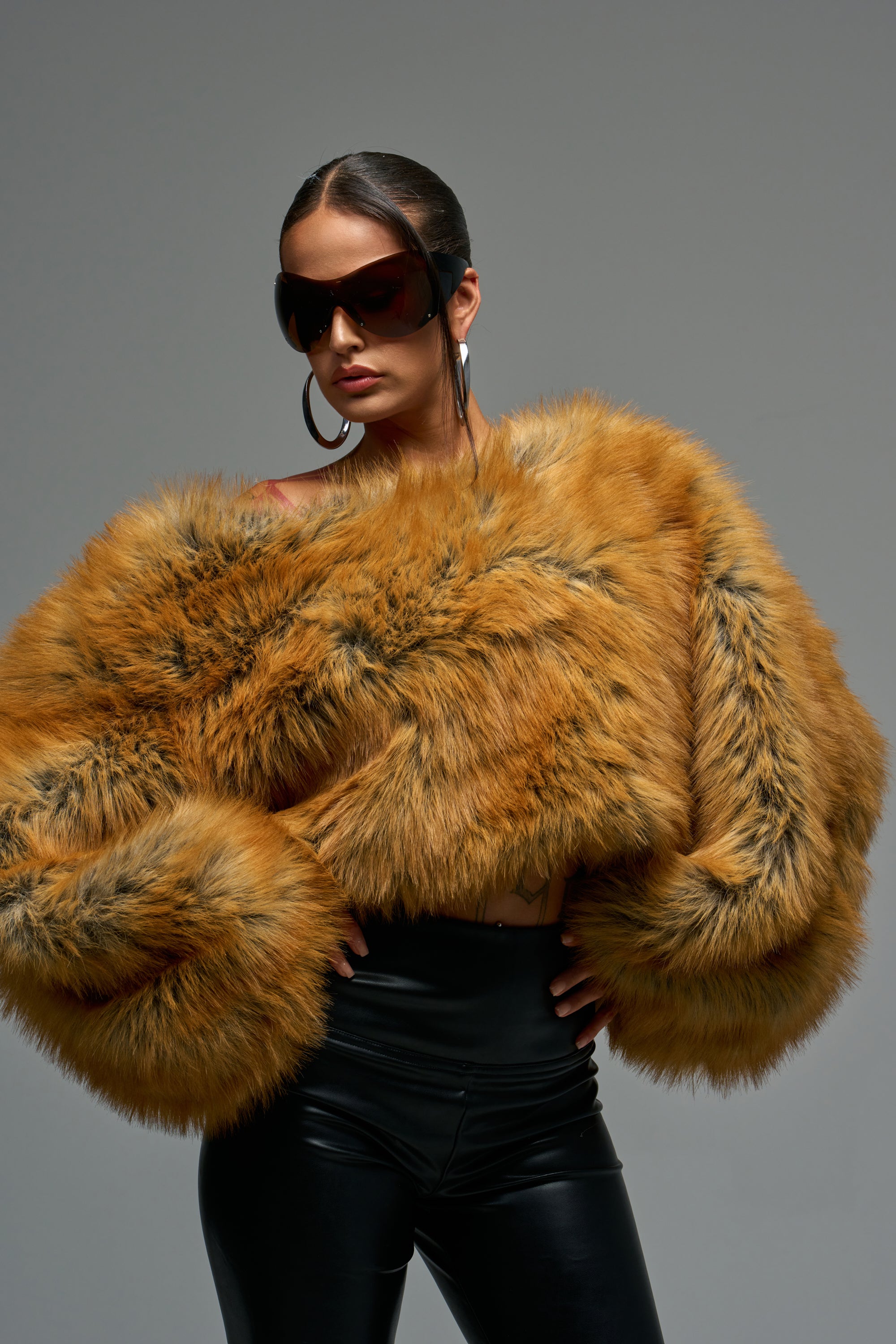 A person poses confidently in the SNOW DAY FAUX FUR SWEATER, oversized sunglasses, large hoop earrings, and black leather pants—epitomizing après-chic cold weather style against a plain gray background.