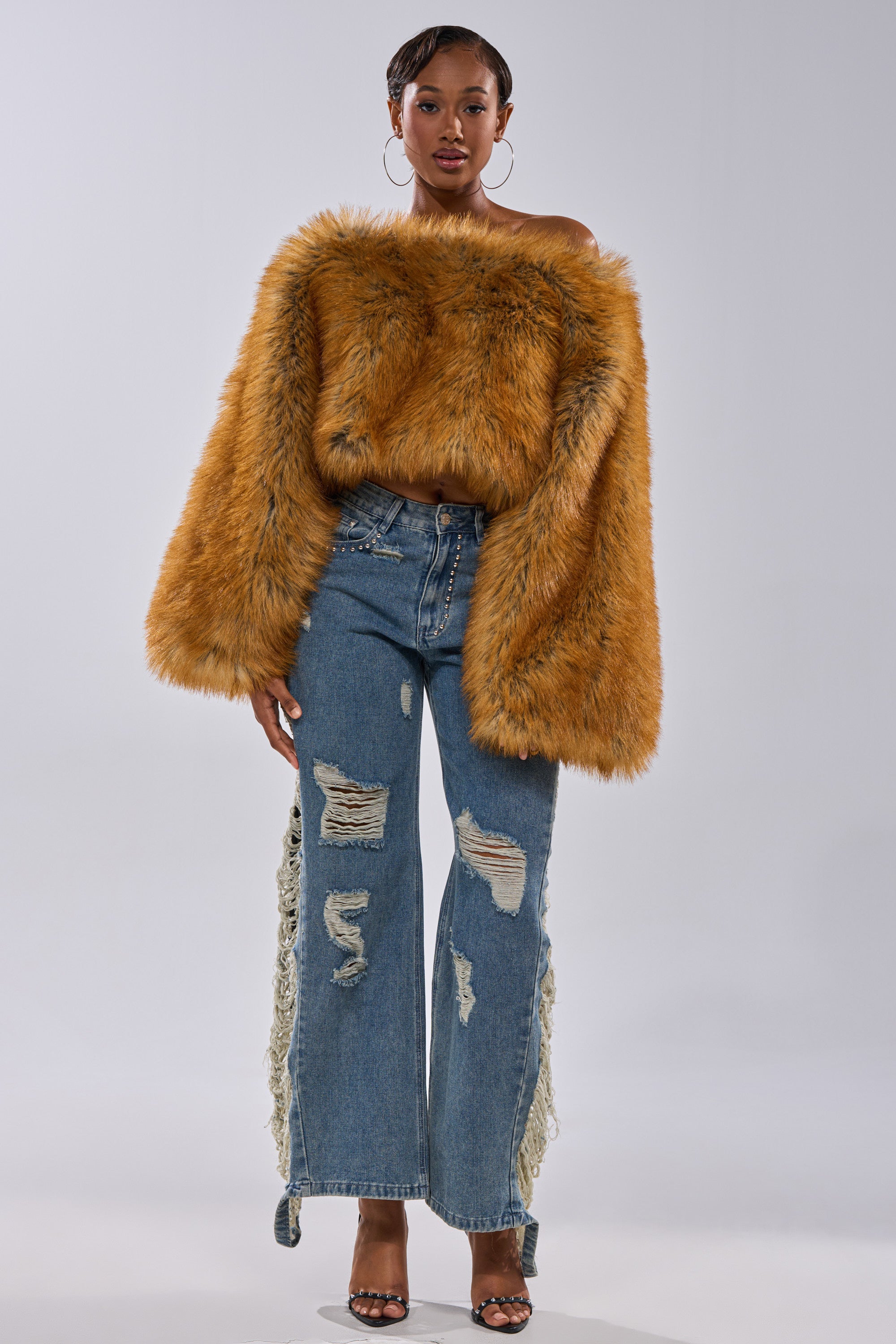 A woman faces forward in the SNOW DAY FAUX FUR SWEATER by AKIRA Label, featuring a fluffy, off-the-shoulder orange design, styled with distressed wide-leg blue jeans and black high-heeled sandals against a plain white background.
