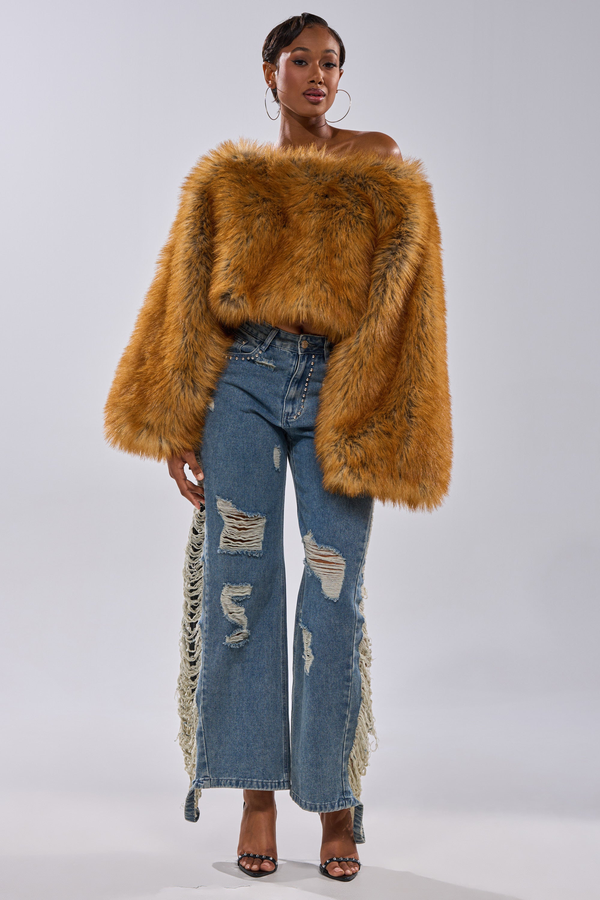 A woman exudes bold winter-chic style in the SNOW DAY FAUX FUR SWEATER, an off-the-shoulder, oversized orange top paired with distressed blue lace-up jeans and black high-heeled sandals against a plain background.