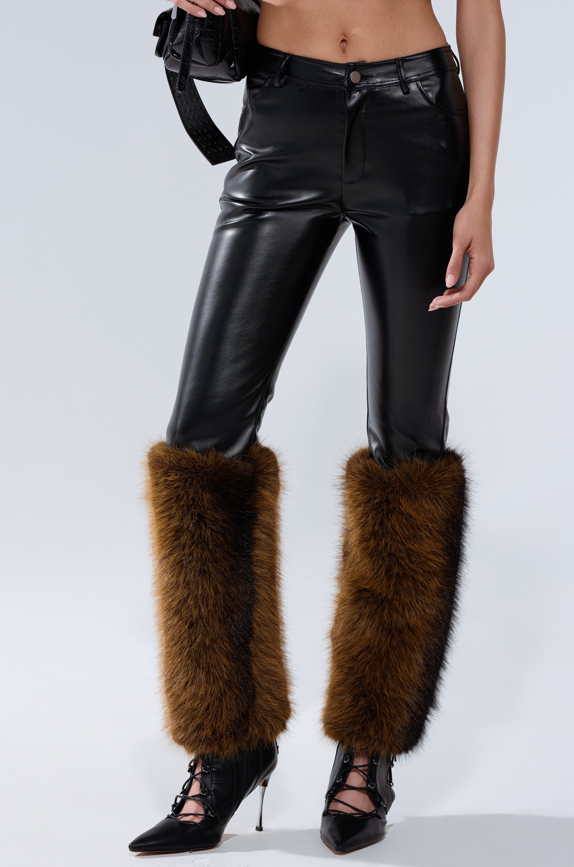 A person models the BABY ITS COLD OUTSIDE FAUX LEATHER PANT WITH FUR TRIM, paired with brown fur leg warmers, black lace-up heels, and a black textured handbag, shown waist down against a light background.