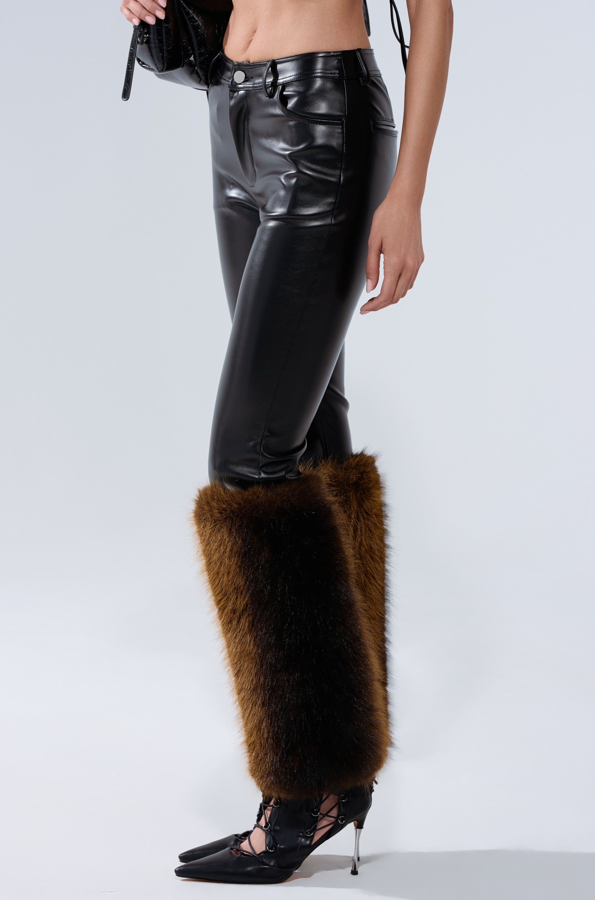 A person models the BABY ITS COLD OUTSIDE FAUX LEATHER PANT WITH FUR TRIM—shiny black high-rise faux leather pants with brown fur trim—styled with black high-heeled shoes and a black handbag against a plain light background.