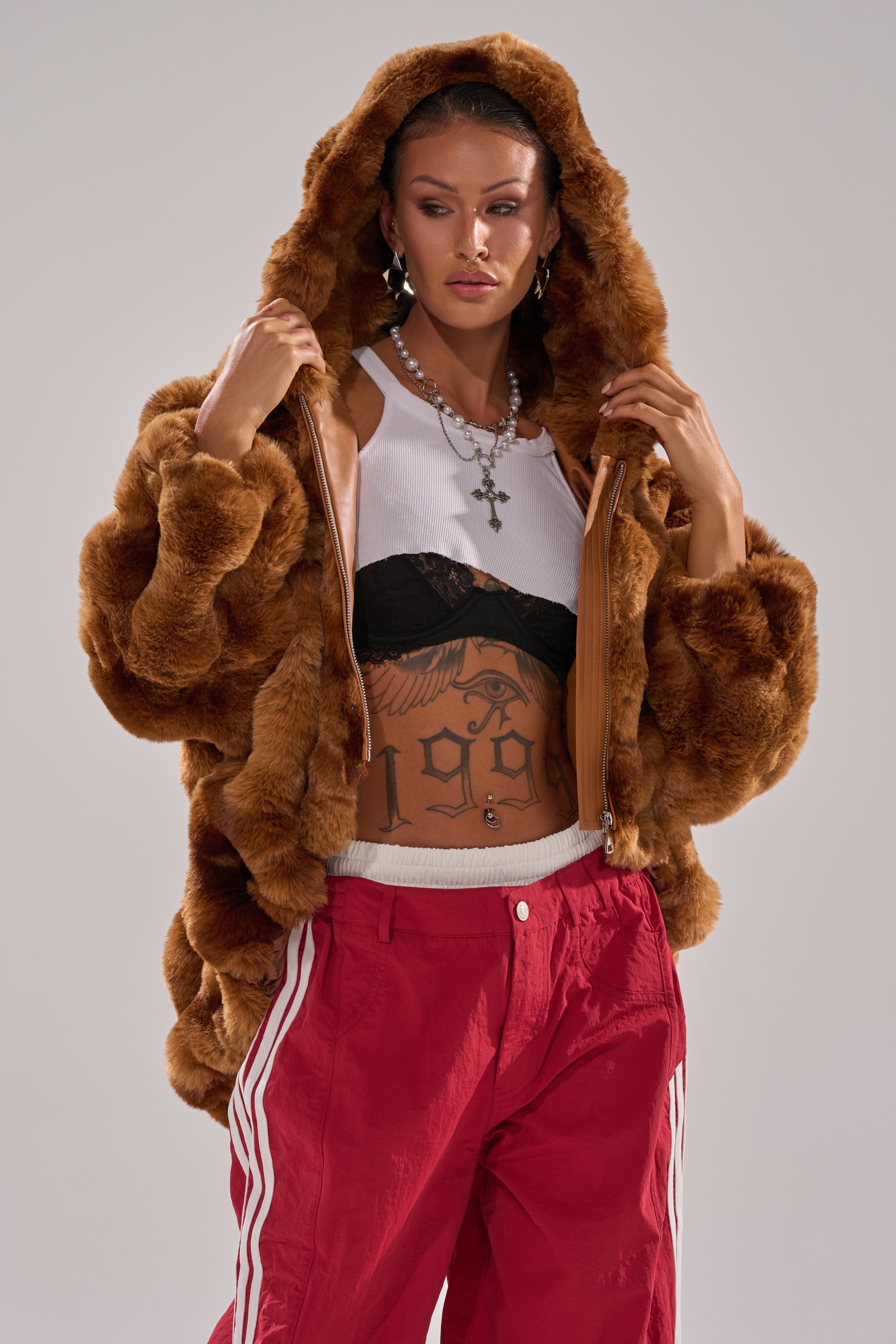Someone stands against a plain background in the LOOK AT ME NOW HOODED FUR BOMBER IN BROWN, paired with a white cropped tank top, black bralette, red track pants with white stripes, and layered necklaces. 0
