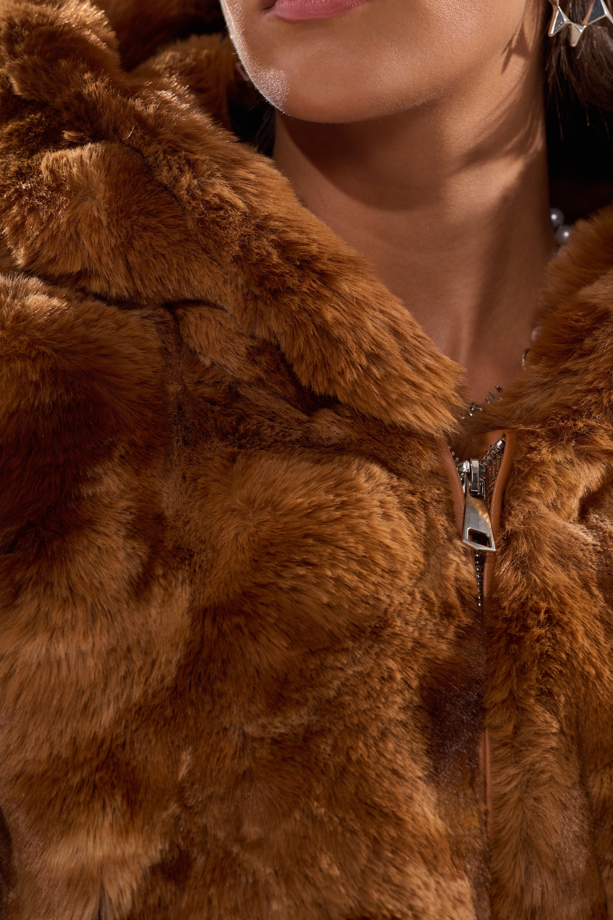 Close-up of a person wearing the LOOK AT ME NOW HOODED FUR BOMBER IN BROWN, featuring soft faux fur, a visible zipper, and a star-shaped earring. Their neck and lips are partially shown. 10