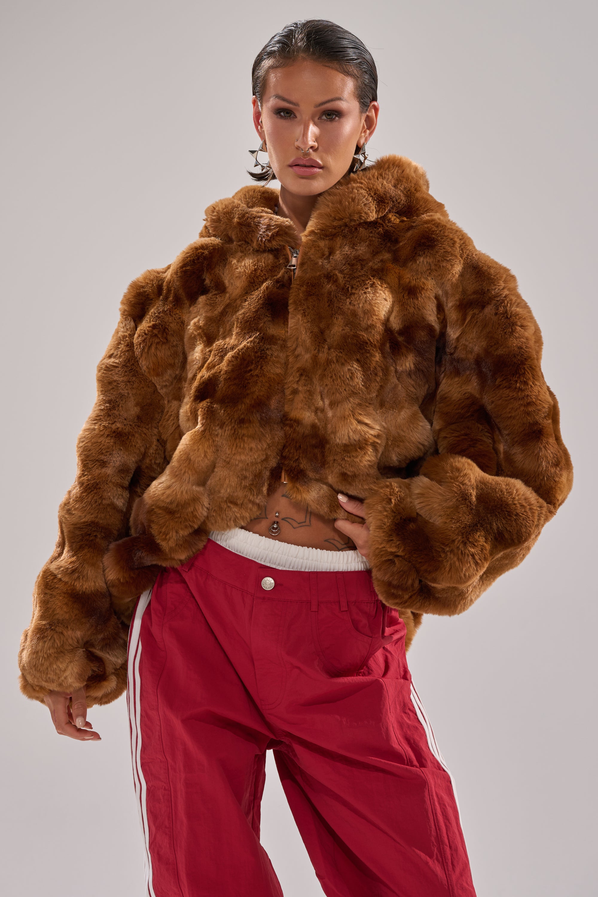 A person wears the LOOK AT ME NOW HOODED FUR BOMBER IN BROWN and red athletic pants with white stripes, standing confidently against a plain background with slicked-back hair and visible piercings. 2