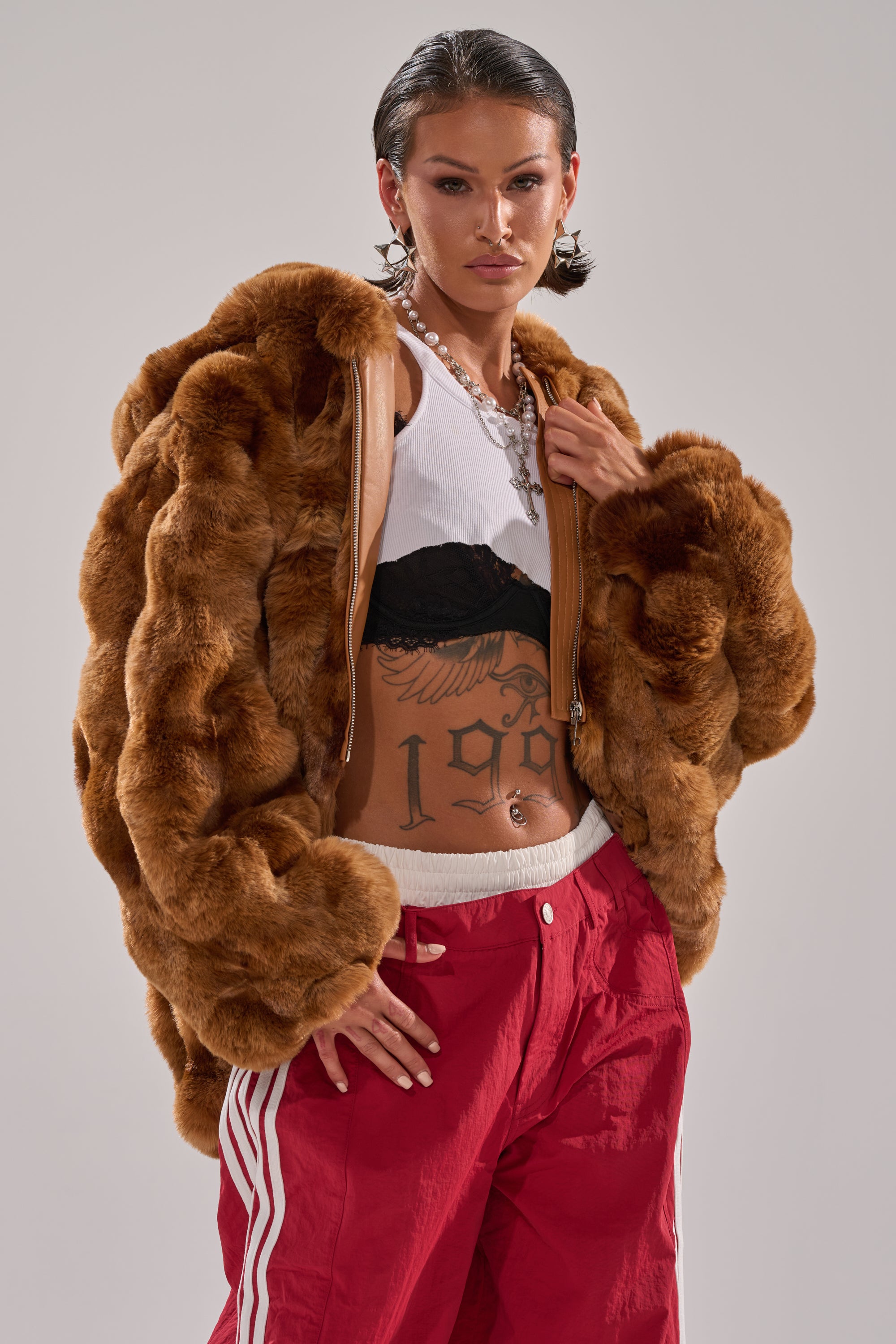 Wearing the LOOK AT ME NOW HOODED FUR BOMBER IN BROWN, white crop top layered with a black bralette, red track pants with white stripes, and silver jewelry, the model poses confidently against a plain background. 3
