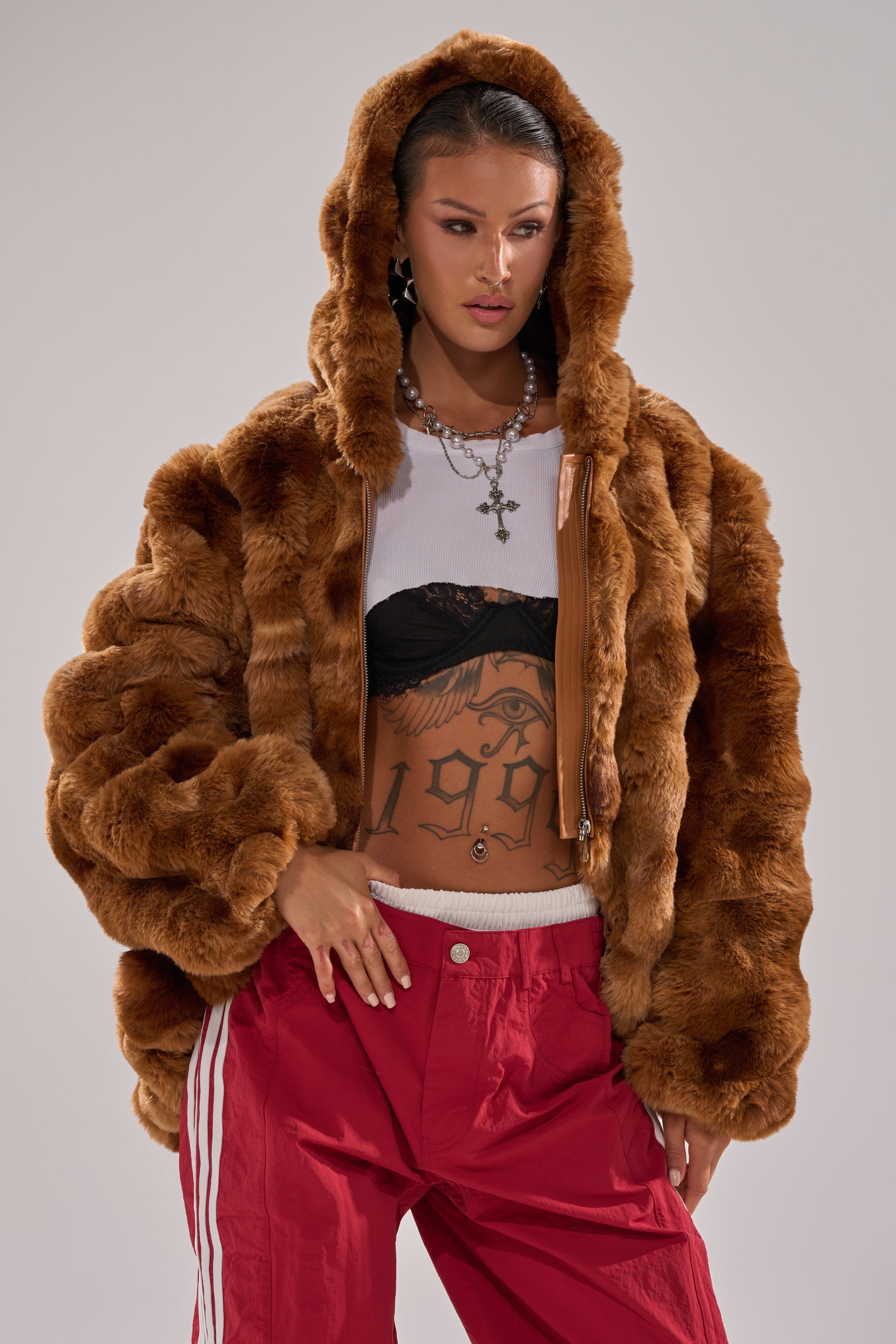 A woman poses confidently in the LOOK AT ME NOW HOODED FUR BOMBER IN BROWN, paired with red striped track pants, a white crop top over a black bra, layered necklaces, and shows off a bold "1990" tattoo on her torso. 1