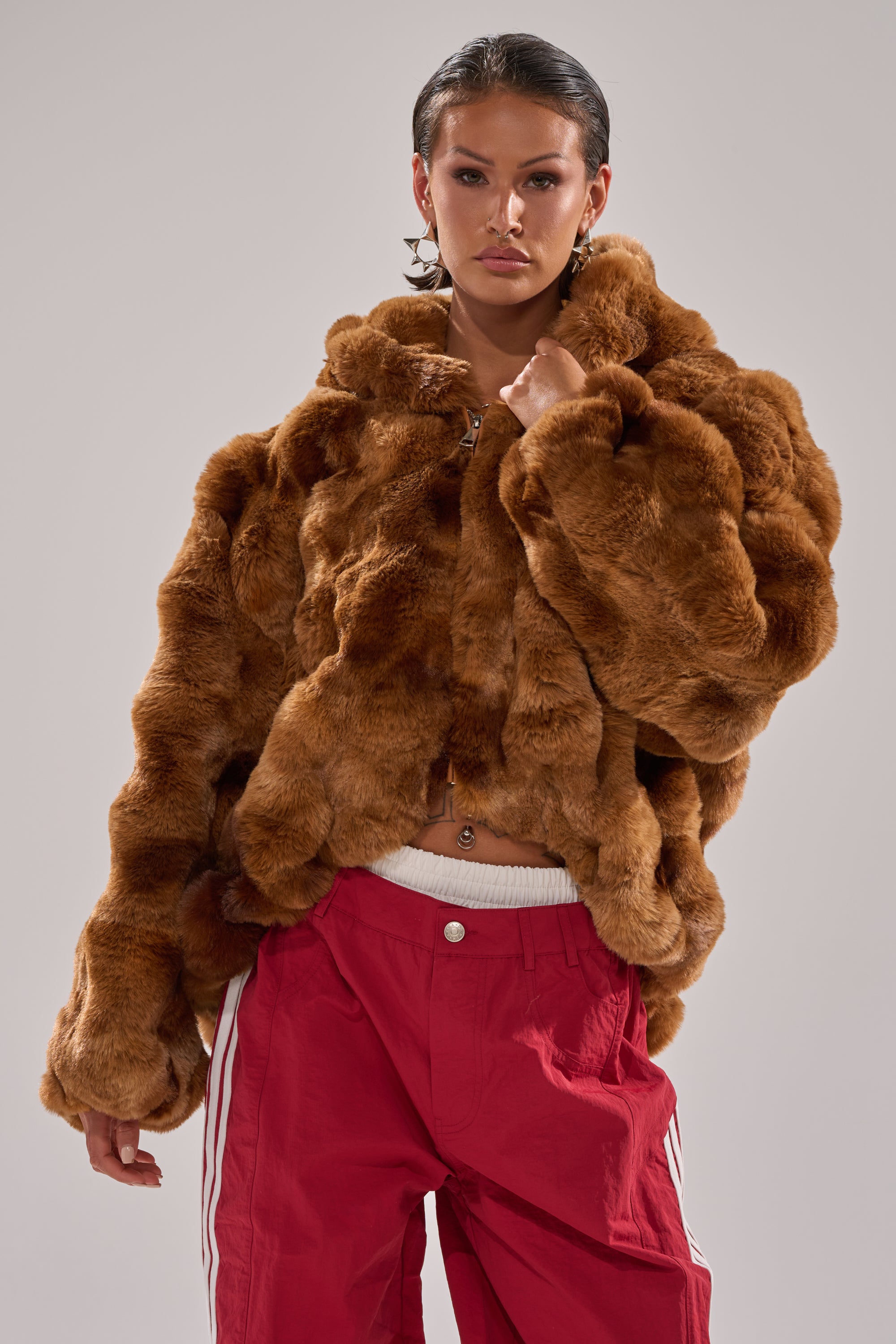 A person stands confidently against a plain light gray background, wearing the LOOK AT ME NOW HOODED FUR BOMBER IN BROWN paired with red track pants featuring white stripes. 4