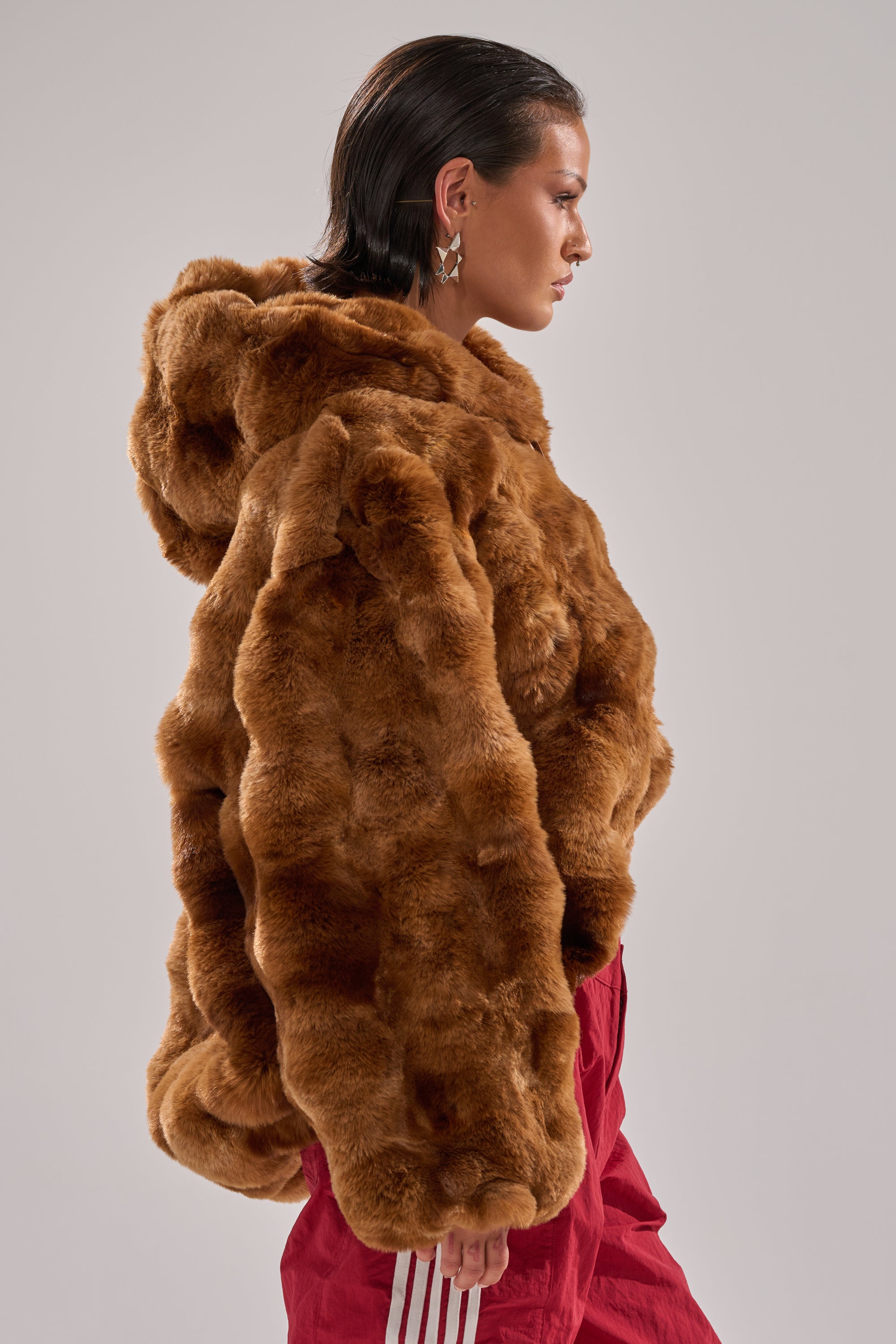 A person with slicked-back dark hair wears the LOOK AT ME NOW HOODED FUR BOMBER IN BROWN, styled in plush faux fur, paired with red pants featuring white stripes, standing in profile against a plain light background. 5