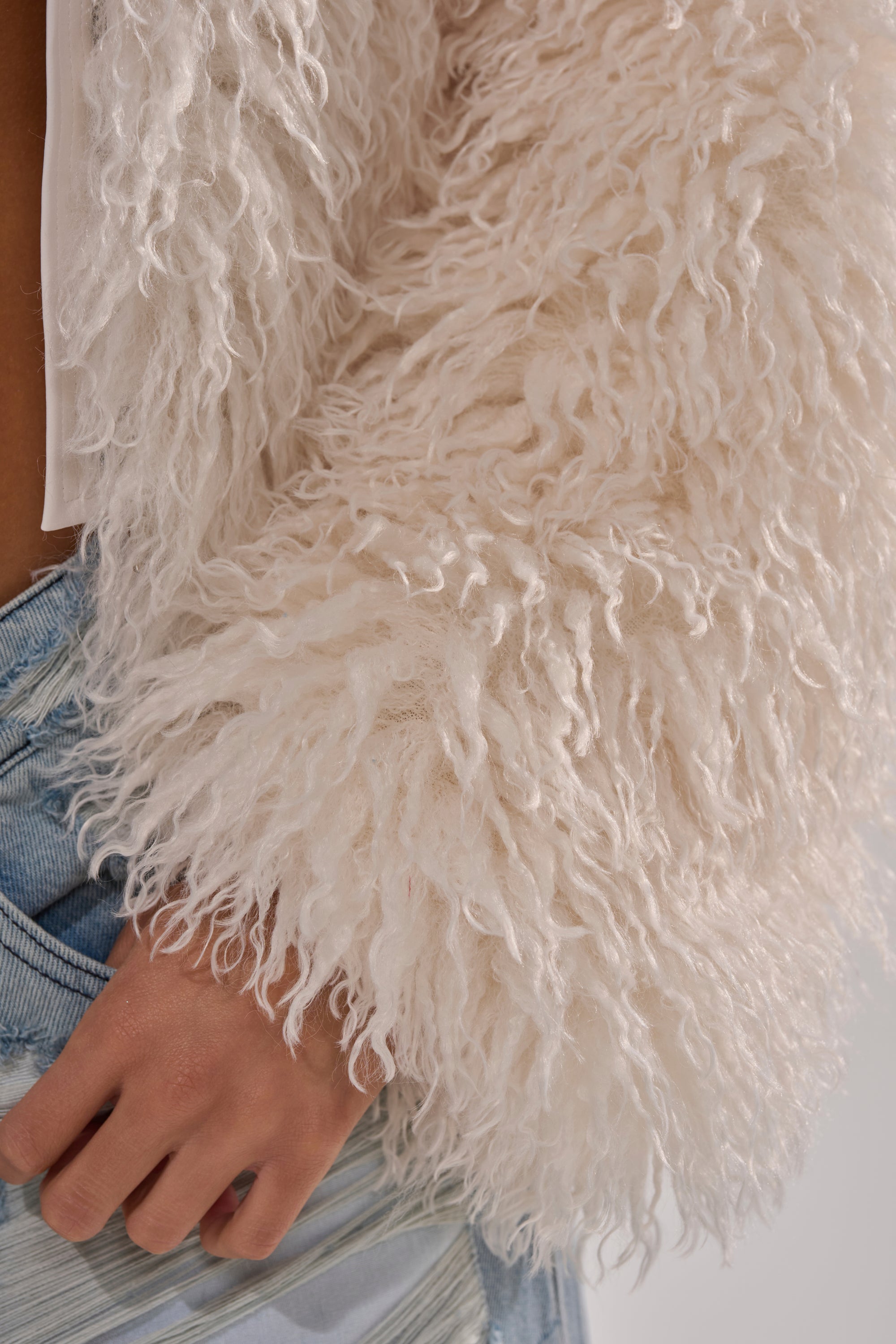 A close-up of a person wearing the LOOK AT ME NOW HOODED FUR BOMBER IN IVORY over a white top and light blue jeans, with their hand in a pocket. 13