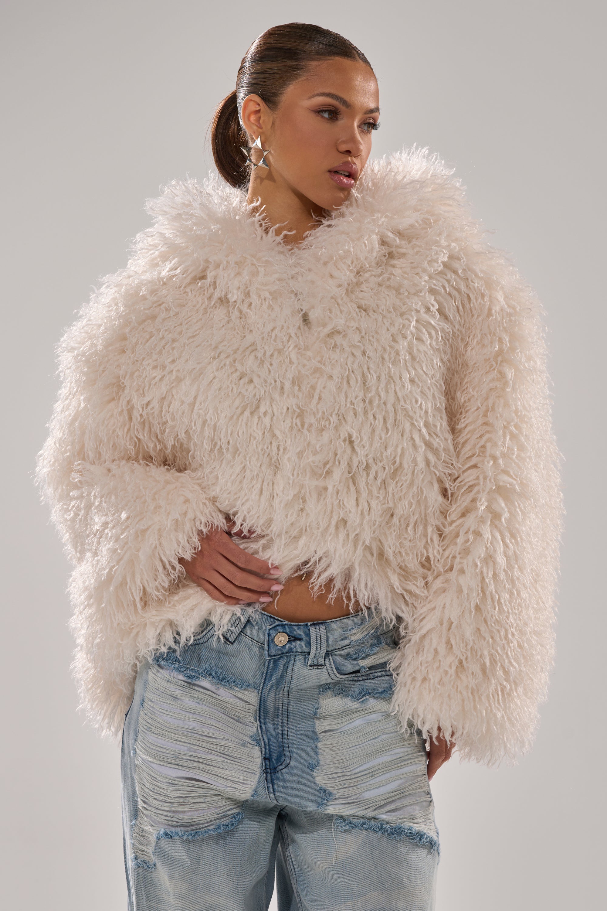 A woman models the LOOK AT ME NOW HOODED FUR BOMBER IN IVORY with distressed light-wash jeans, standing against a plain background and gazing to the side, her hand resting on her hip. 5