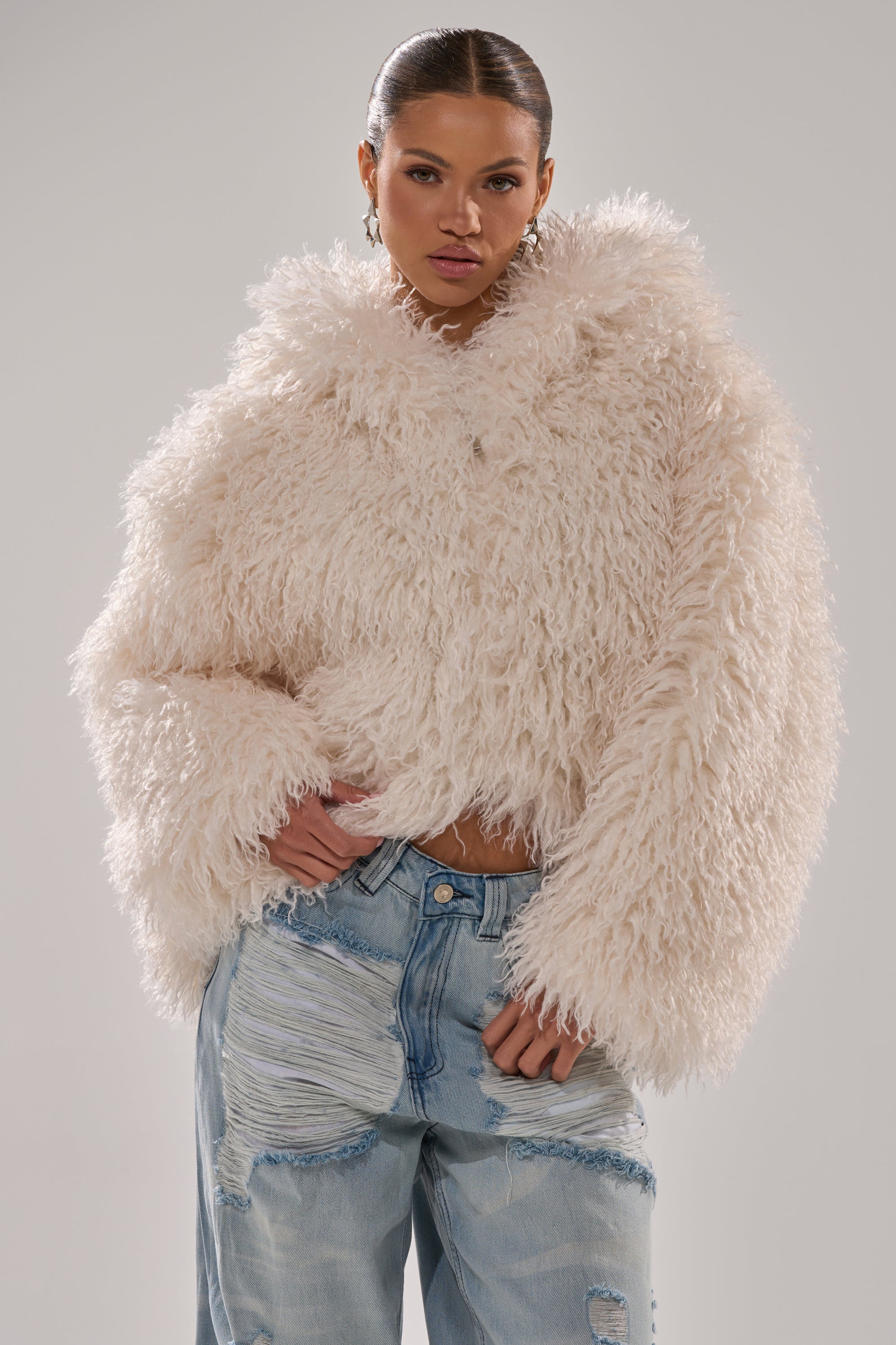 A woman stands confidently against a plain gray background wearing the LOOK AT ME NOW HOODED FUR BOMBER IN IVORY and ripped light blue jeans. 6