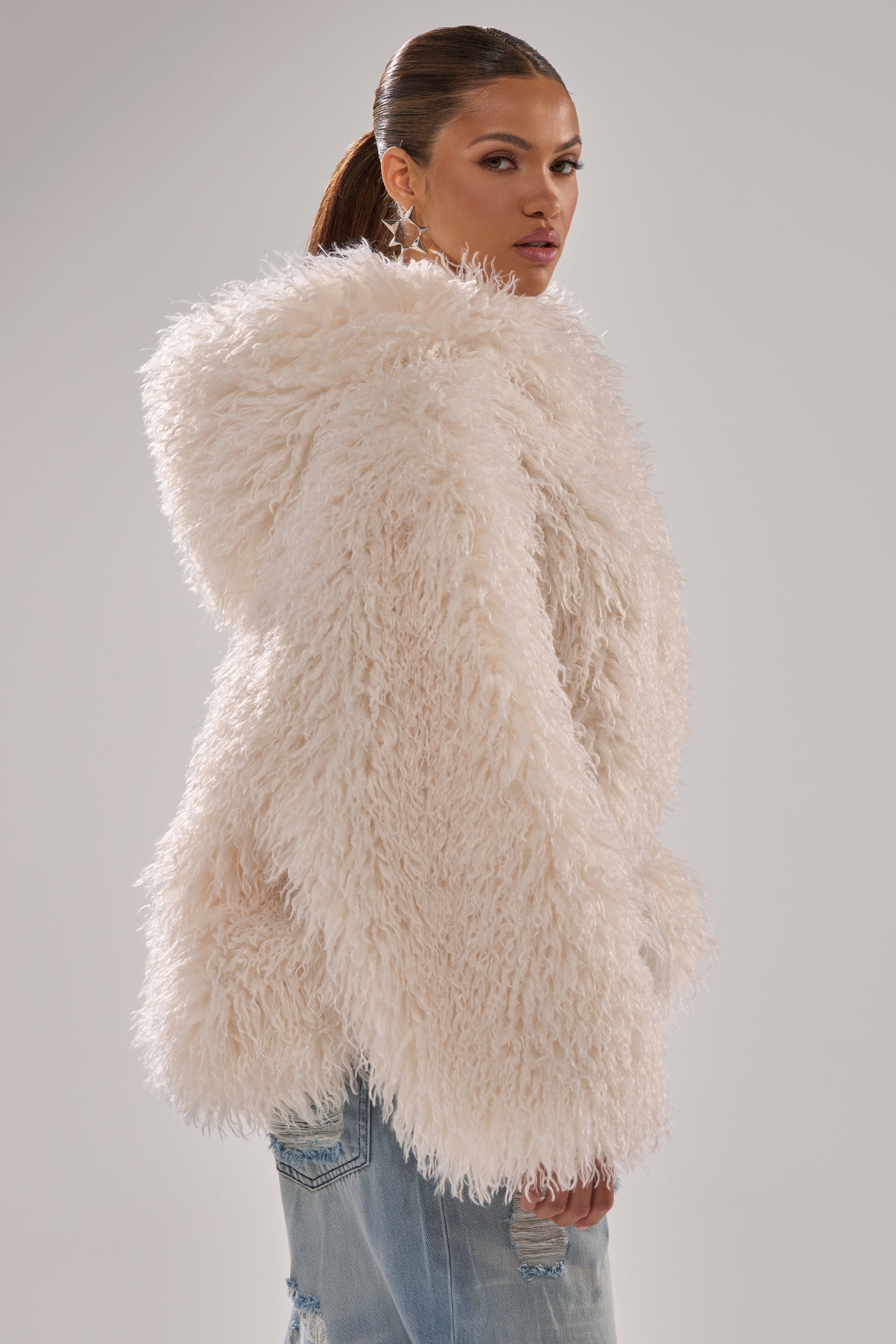 A woman with slicked-back hair models the LOOK AT ME NOW HOODED FUR BOMBER IN IVORY and light blue ripped jeans, standing against a plain light gray background and looking over her shoulder. 8