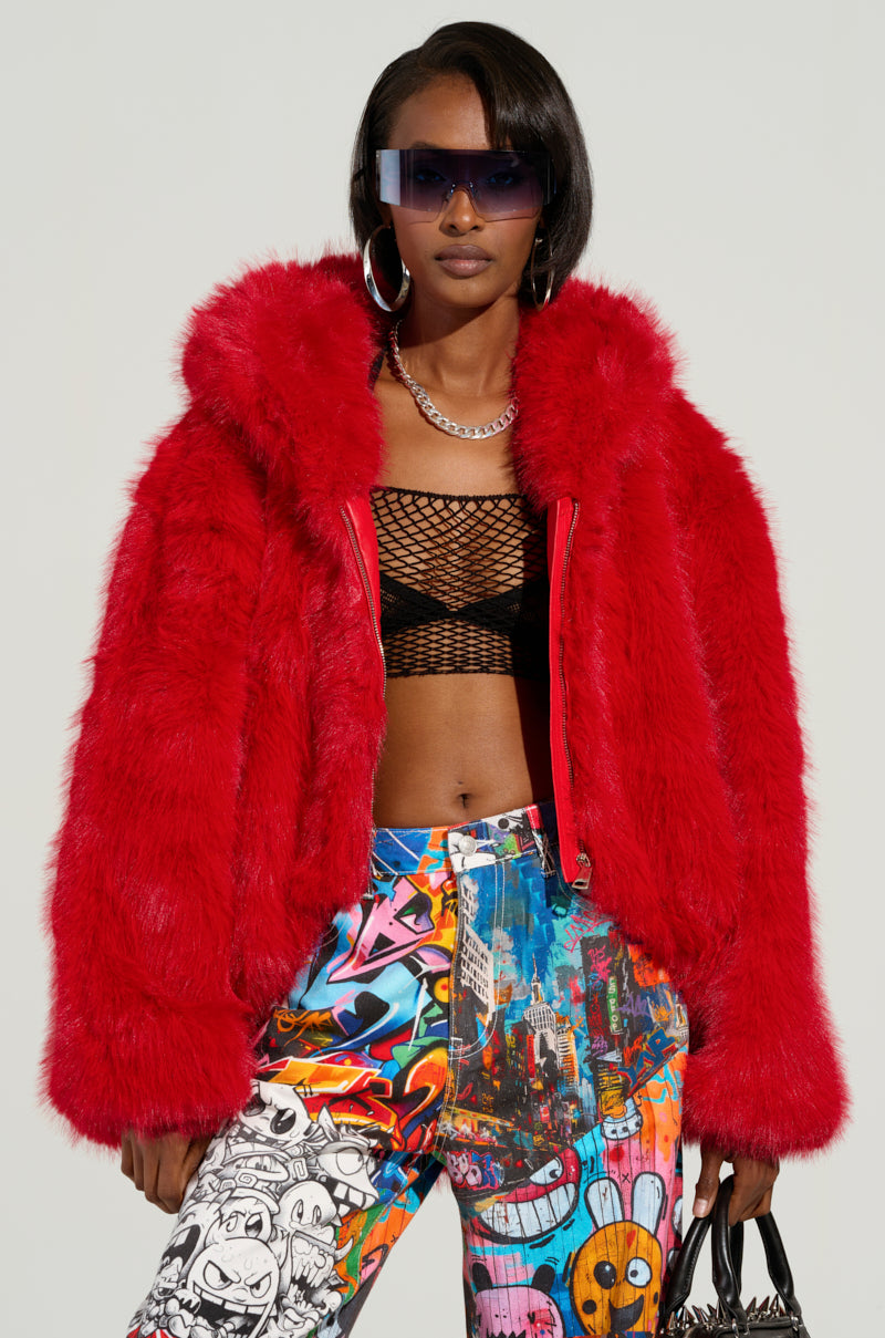 A woman flaunts the LOOK AT ME NOW HOODED FUR BOMBER in a vibrant red color from the AKIRA Label, paired with a black mesh top and colorful graffiti-patterned pants. She accessorizes with large sunglasses, hoop earrings, and a chain necklace while holding a stylish handbag against a plain backdrop.