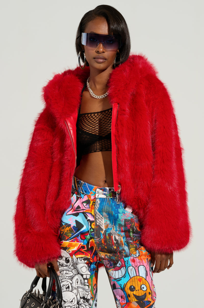 A person is dressed in a vibrant red LOOK AT ME NOW HOODED FUR BOMBER from the AKIRA Label, paired with a black mesh top and graffiti-patterned pants. They complete their look with large sunglasses, a chunky necklace, and carry a studded black bag against a plain light gray background.