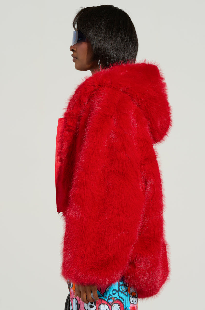 A person with short hair and sunglasses dons a striking LOOK AT ME NOW Hooded Fur Bomber in vibrant red faux fur. The jacket stands out against their colorful pants adorned with abstract patterns. They are turned slightly to the side against a neutral backdrop, showcasing an audacious sense of style.