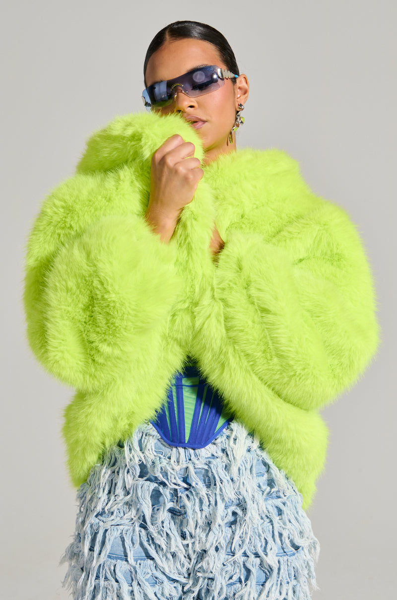 A person poses in a LANA Hooded Faux Fur Coat in lime, paired with stylish blue sunglasses, dangling earrings, and a blue corset. The skirt is crafted from shredded denim fabric. The neutral background highlights the vibrant ensemble, ensuring the stand-out beauty of the outfit doesn't go unnoticed.