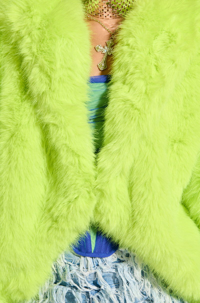 Close-up of a person sporting the LANA Hooded Faux Fur Coat in Lime from the AKIRA Label. They wear a gold cross necklace layered over a blue top, complemented by a garment with a distressed, shredded texture.