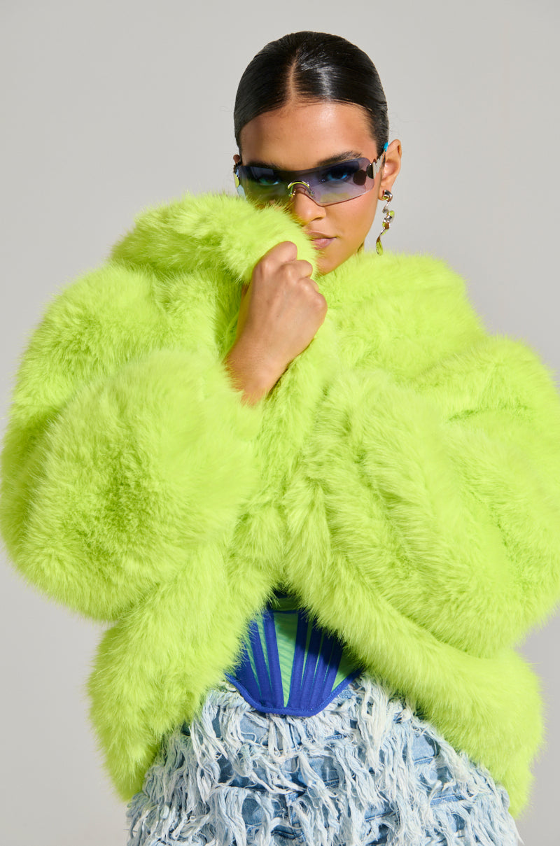 A person wearing the vibrant LANA Hooded Faux Fur Coat in Lime from the AKIRA Label, blue-tinted sunglasses, and a textured skirt looks confidently at the camera. The background is a plain gray, highlighting the bold, fashionable outfit.