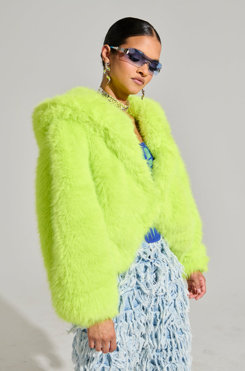 A person wearing a vibrant LANA Hooded Faux Fur Coat in Lime from the AKIRA Label and stylish sunglasses. They have sleek black hair adorned with large, dangling earrings. The lower outfit features an intricate, textured pattern in light blue. They pose confidently.
