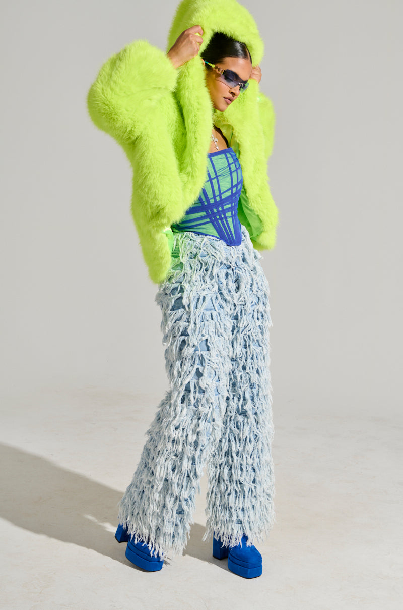 A person wearing the LANA HOODED FAUX FUR COAT IN LIME from the AKIRA Label, a vibrant blue and green bodysuit, and fluffy light blue textured pants. They accessorize with dark sunglasses and blue platform shoes, striking a confident pose against a plain background.