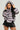 A person poses confidently in an oversized silhouette, wearing the MINA FAUX FUR BOMBER, a fluffy grey hooded coat with stripes and a ribbed waistband. Paired with this is a black pleated skirt featuring metal accents. The individual, with long black hair, stands gracefully against a neutral background.