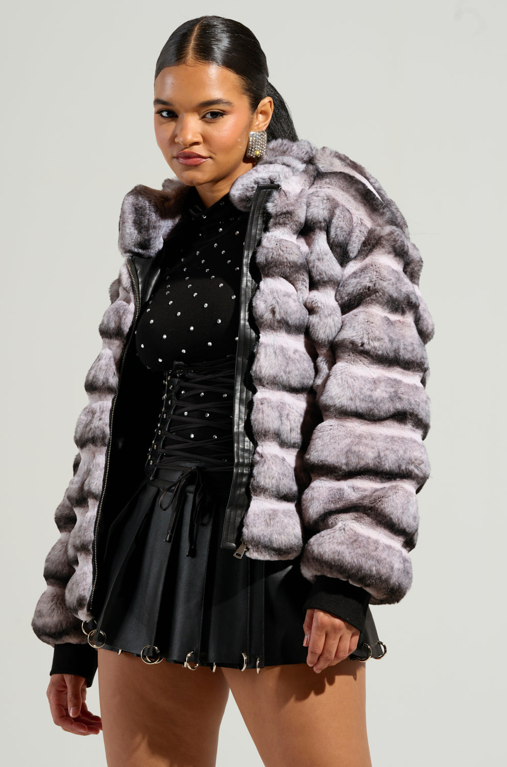 Sporting the MINA FAUX FUR BOMBER in a luxurious silver-gray, a woman pairs it with a black high-neck top adorned with white polka dots and a sleek black skirt. Her hair is elegantly pulled back as she gazes confidently at the camera, the bomber's oversized silhouette amplifying her chic elegance.