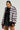 Sporting the MINA FAUX FUR BOMBER in a luxurious silver-gray, a woman pairs it with a black high-neck top adorned with white polka dots and a sleek black skirt. Her hair is elegantly pulled back as she gazes confidently at the camera, the bomber's oversized silhouette amplifying her chic elegance.