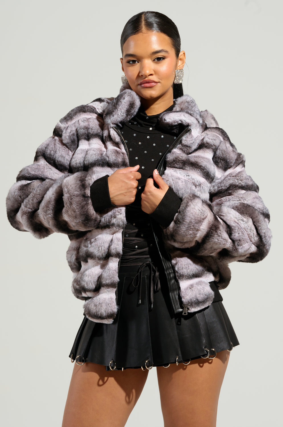A woman poses confidently in the MINA FAUX FUR BOMBER, showcasing an oversized silhouette over a black patterned top and black mini skirt. Her slicked-back hair complements her large earrings, all set against a plain, light-colored backdrop.