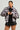 A woman poses confidently in the MINA FAUX FUR BOMBER, showcasing an oversized silhouette over a black patterned top and black mini skirt. Her slicked-back hair complements her large earrings, all set against a plain, light-colored backdrop.
