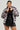 Wearing an oversized silhouette, a person confidently holds the collar of their fluffy MINA FAUX FUR BOMBER in light gray. Underneath, they sport a black shirt with white polka dots paired with a black skirt, looking forward with determination.