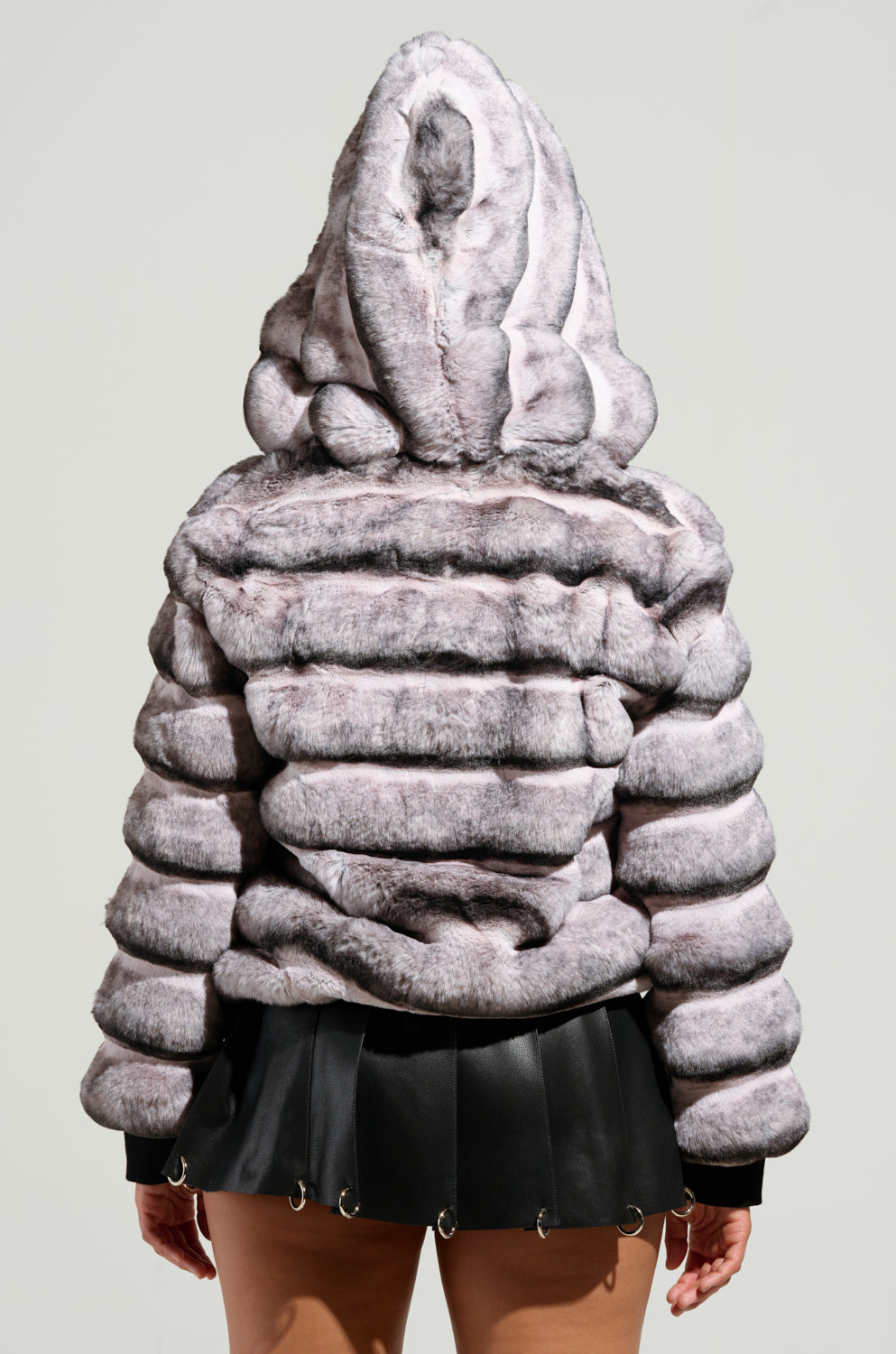 The image features a person from behind wearing the MINA FAUX FUR BOMBER, which has an oversized silhouette with plush, striped faux fur and includes a hood. The jacket is paired with a short black skirt, set against a plain, neutral background.