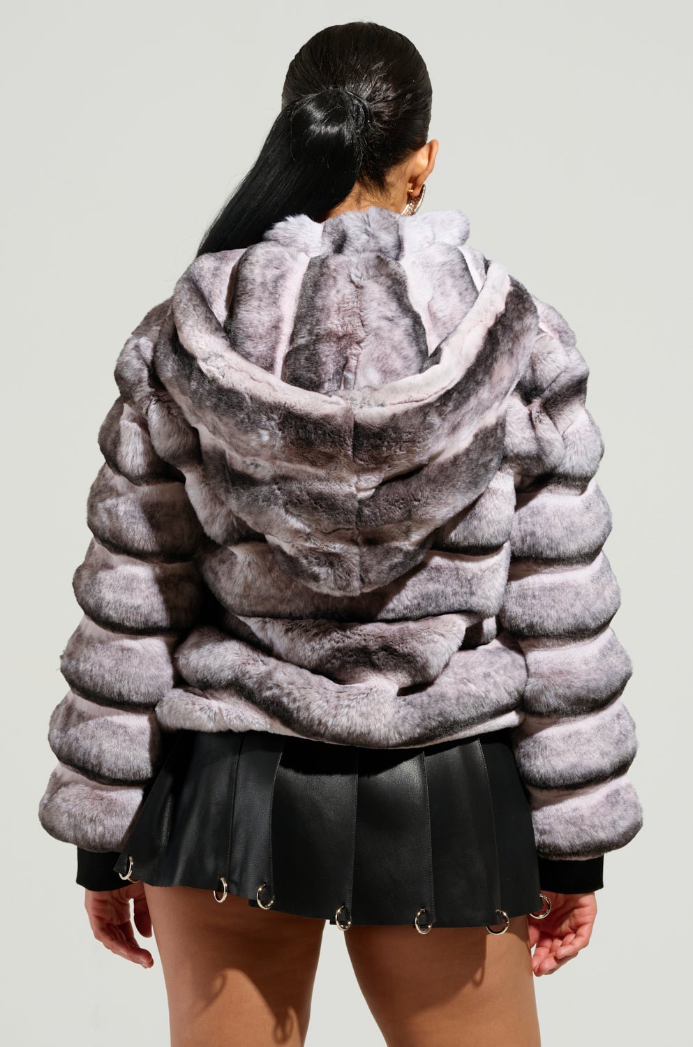 A person with long black hair is seen from the back, wearing the oversized MINA FAUX FUR BOMBER, characterized by its fluffy striped design and hood. They pair it with a short black skirt featuring metal ring accents.