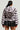 A person with long black hair is seen from the back, wearing the oversized MINA FAUX FUR BOMBER, characterized by its fluffy striped design and hood. They pair it with a short black skirt featuring metal ring accents.